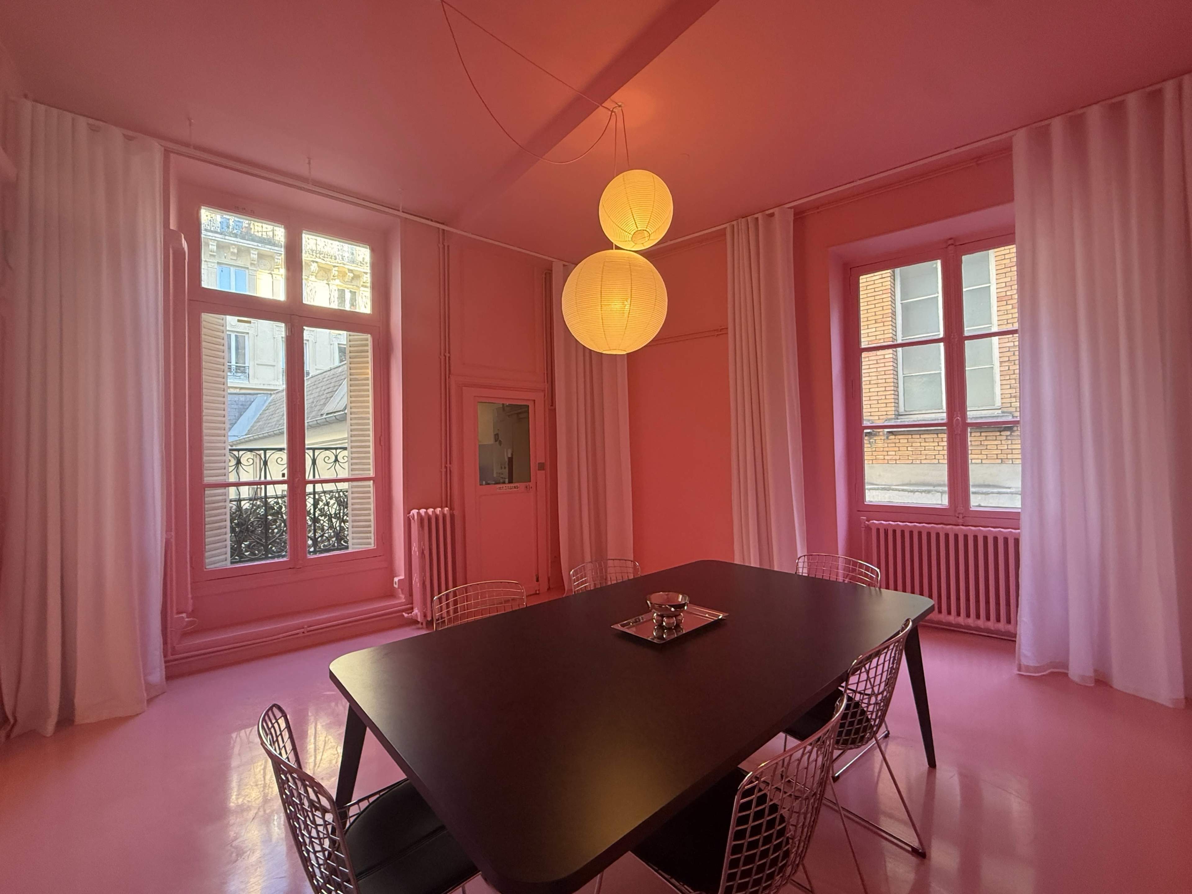 A dining room features pink walls, a large black table surrounded by wire chairs, and two round pendant lights hanging from the ceiling.