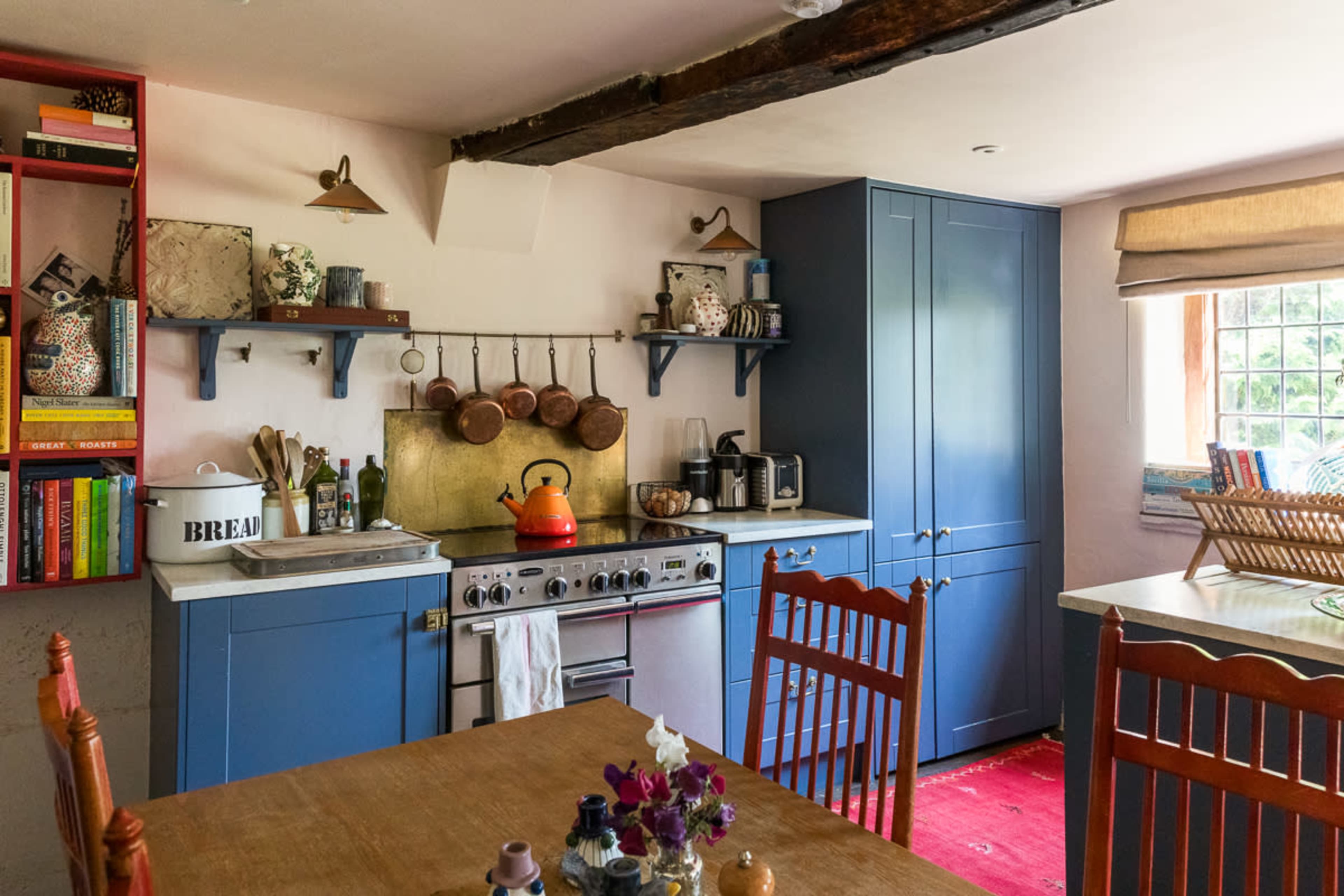 The image shows a kitchen with blue cabinetry, wooden shelves displaying various kitchen items, and a dining table set with flowers.