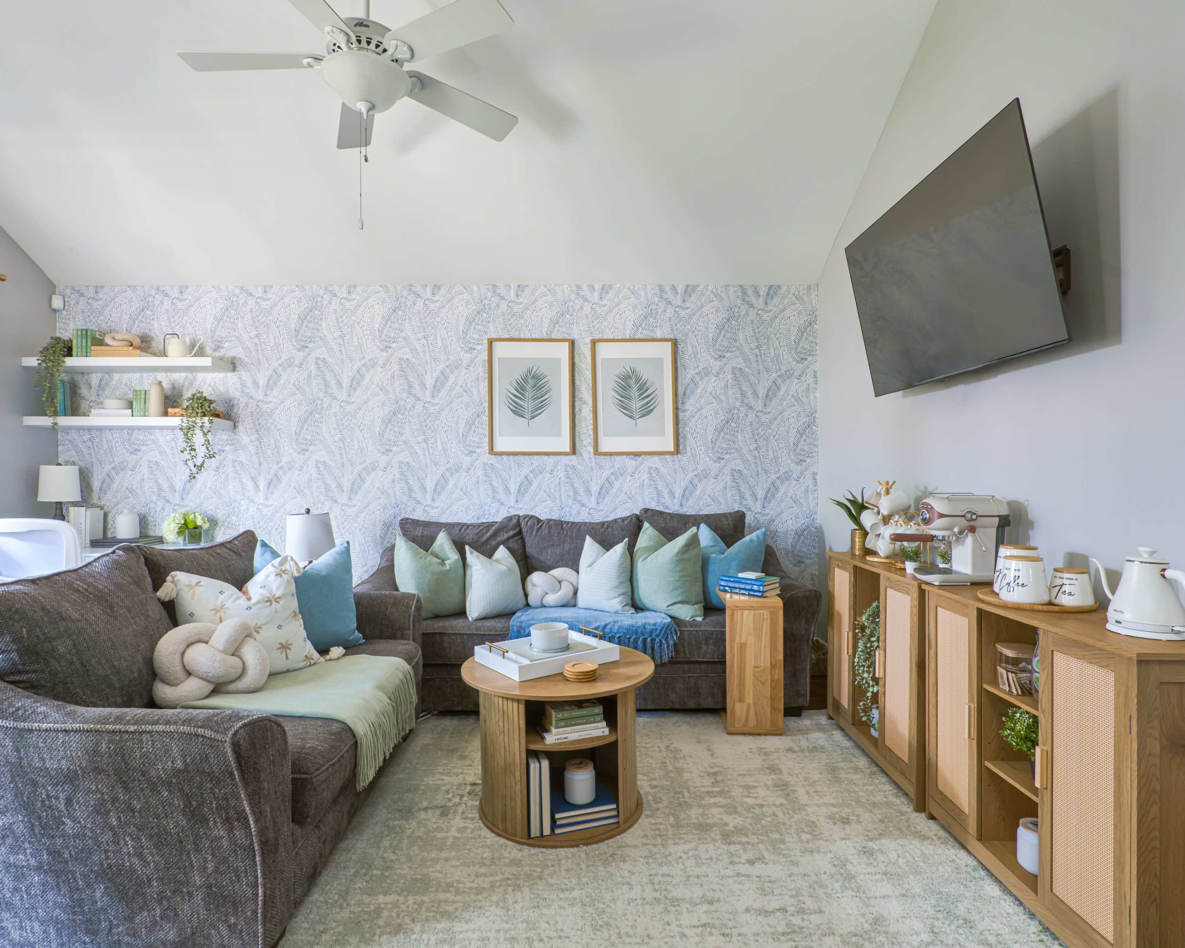 The living room features a gray sectional sofa adorned with decorative pillows, a round wooden coffee table, and a mounted television, all set against a wallpaper with a leaf pattern.