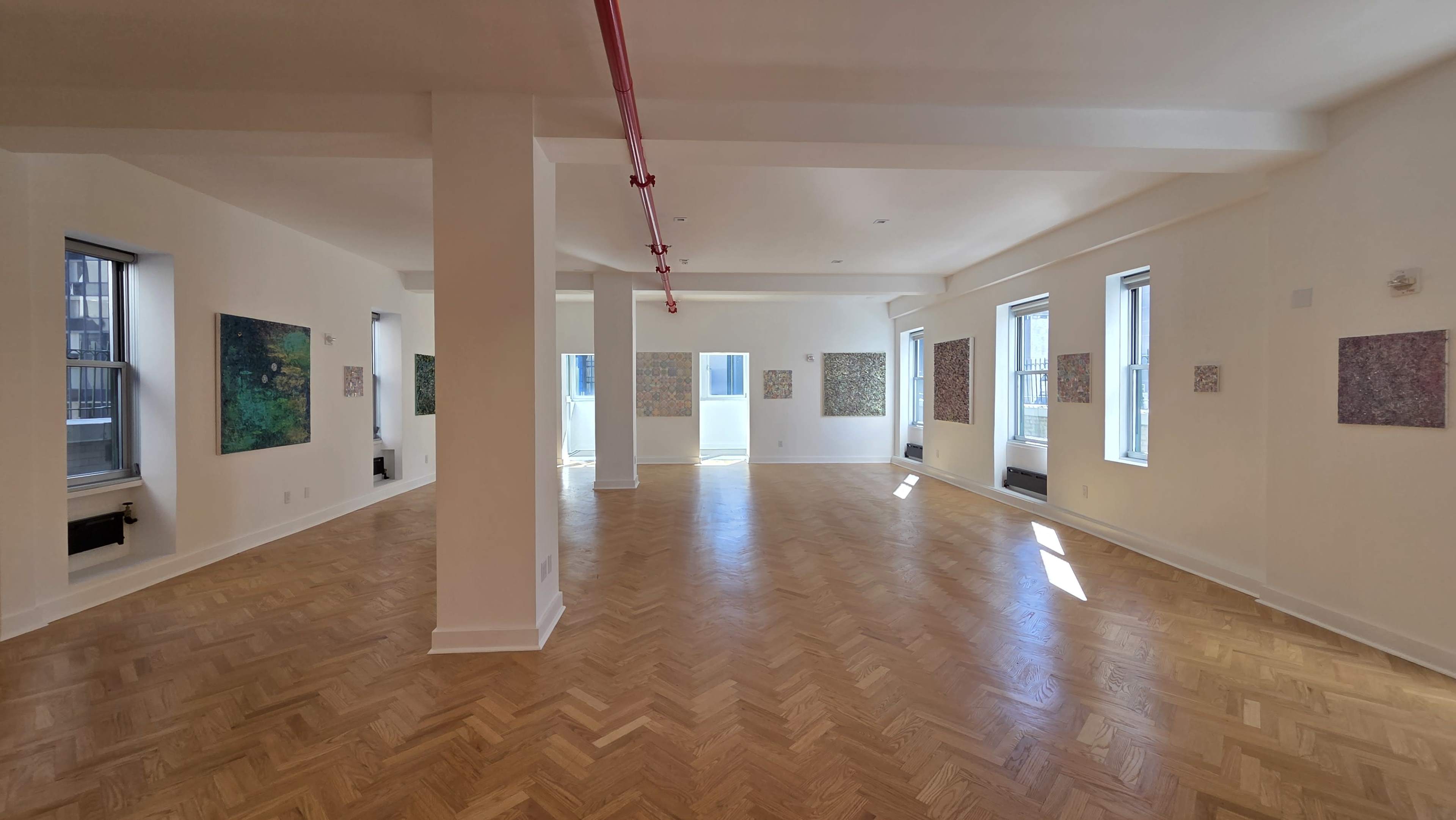 The image shows a spacious, well-lit room with wooden herringbone flooring and several abstract paintings displayed on the walls.