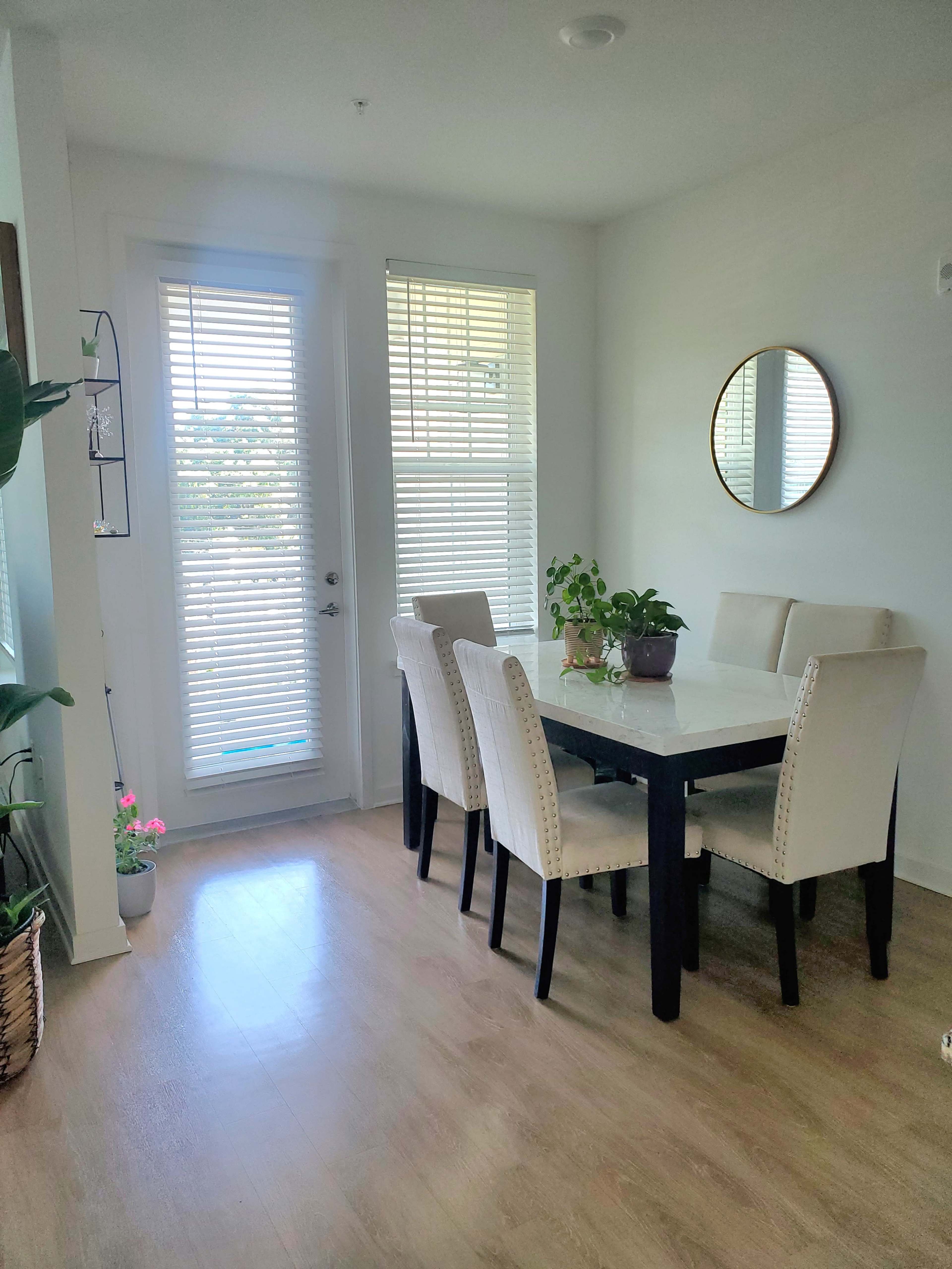 A dining area with a white table, six chairs, a potted plant, and large windows covered with blinds.