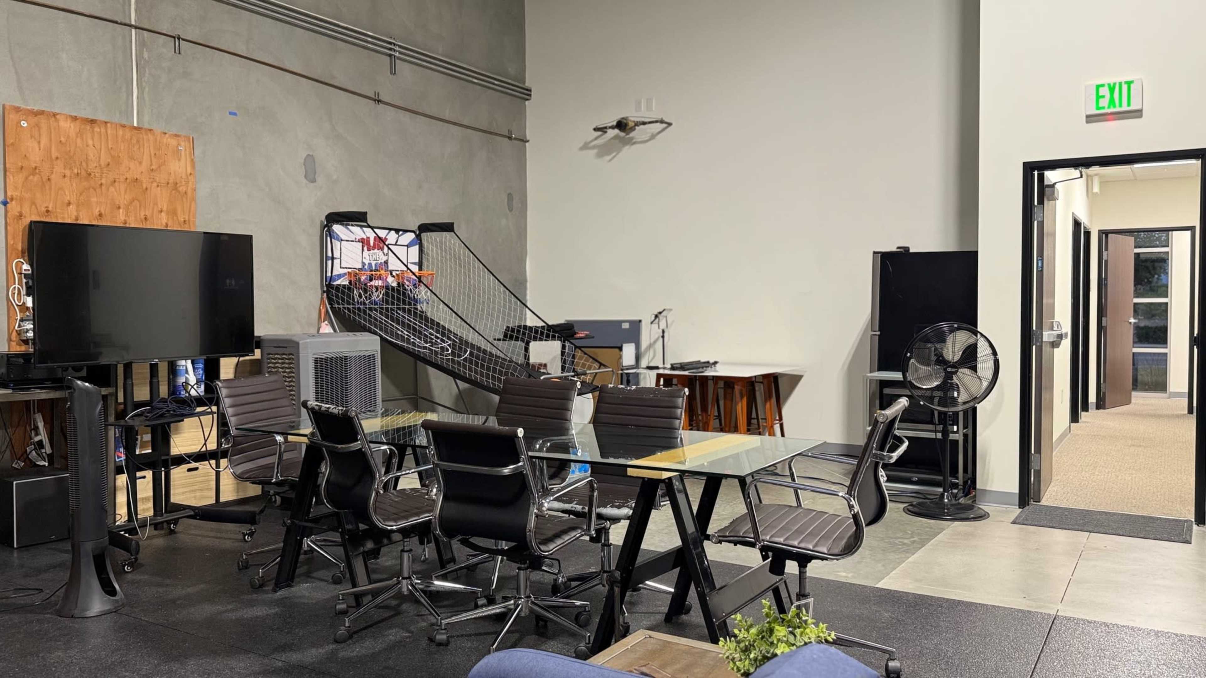 The image shows an office space featuring a conference table with several chairs, a basketball hoop, and a few electronic devices including monitors.
