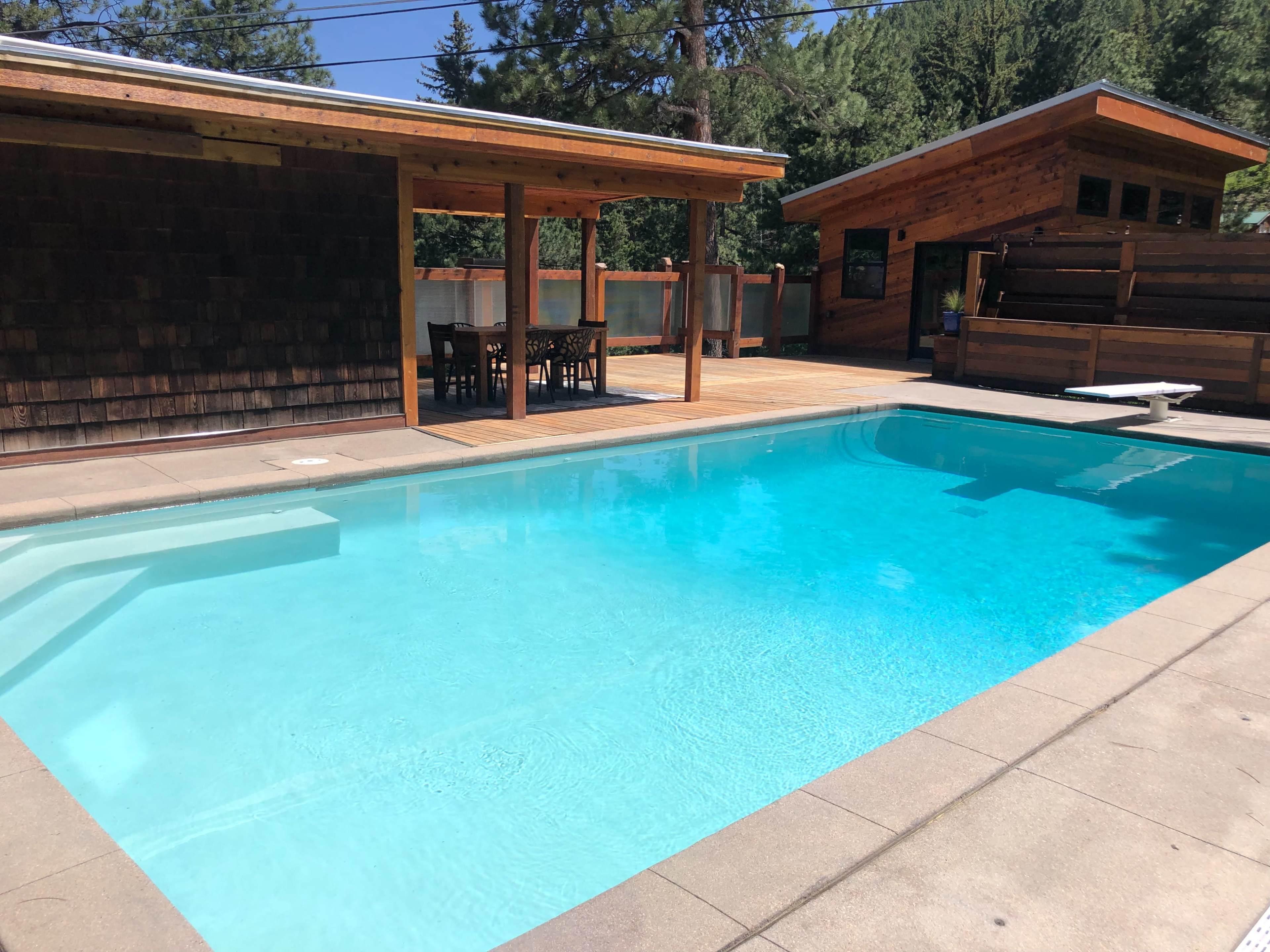 The image shows a clear swimming pool surrounded by wooden structures and a deck area with outdoor seating.