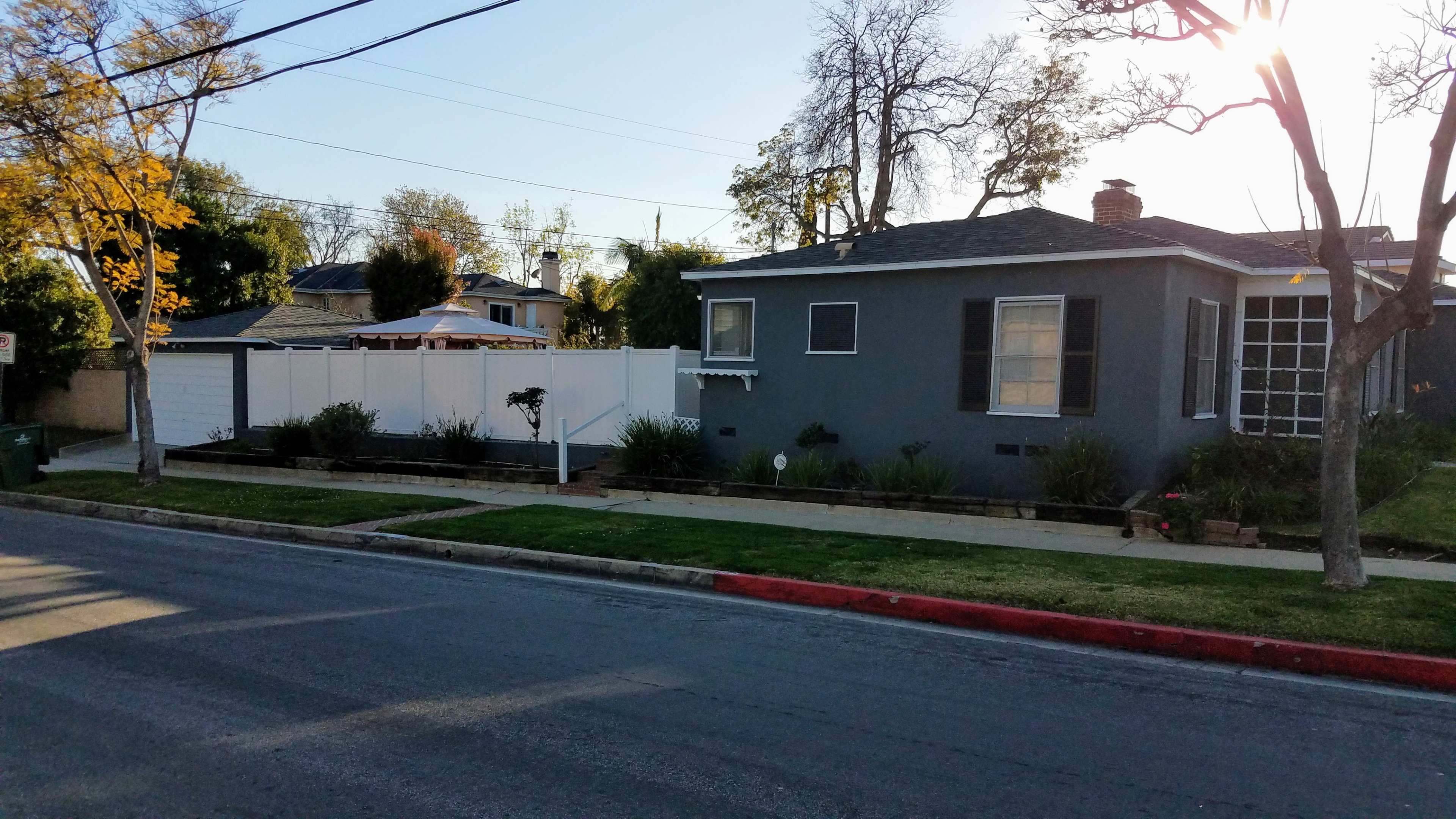 The image shows a single-story gray house with a front yard and a white fence beside a quiet street.