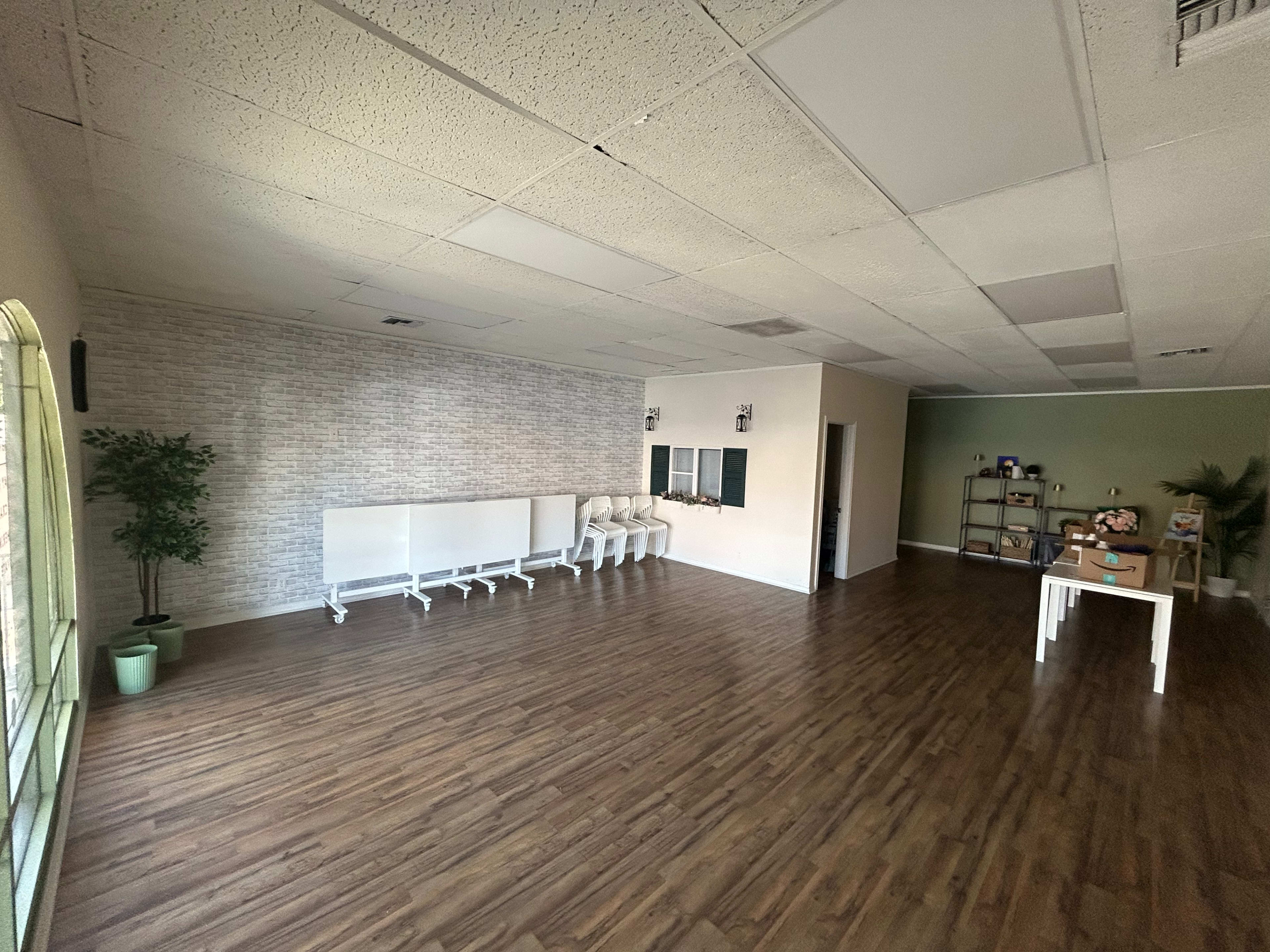 The image shows a spacious, partially furnished room with a light wood floor, a few plants, and a table along with empty chairs lined against one wall.