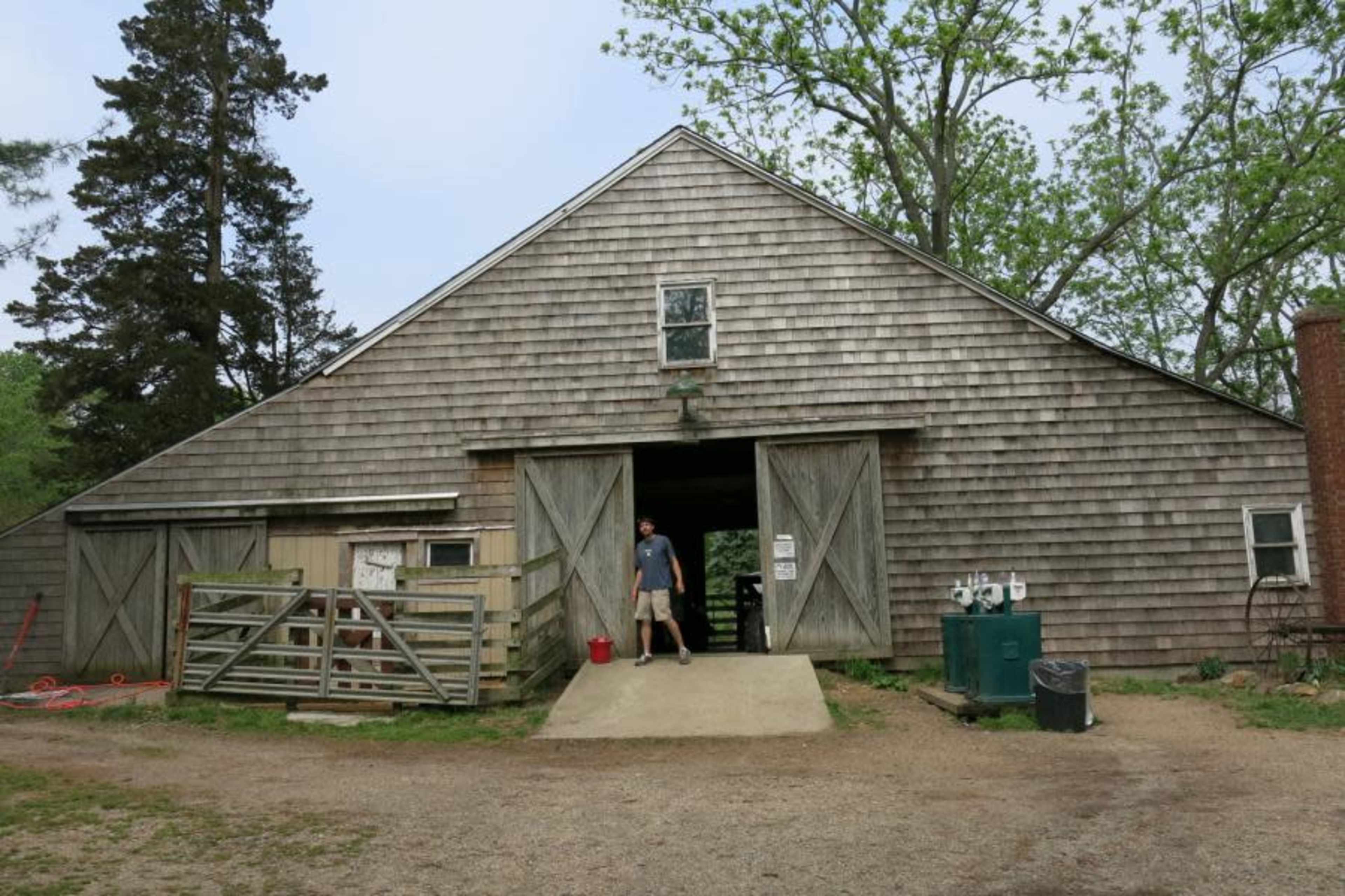 A person stands in front of a large, wooden barn with open double doors and a gravel pathway leading up to it.