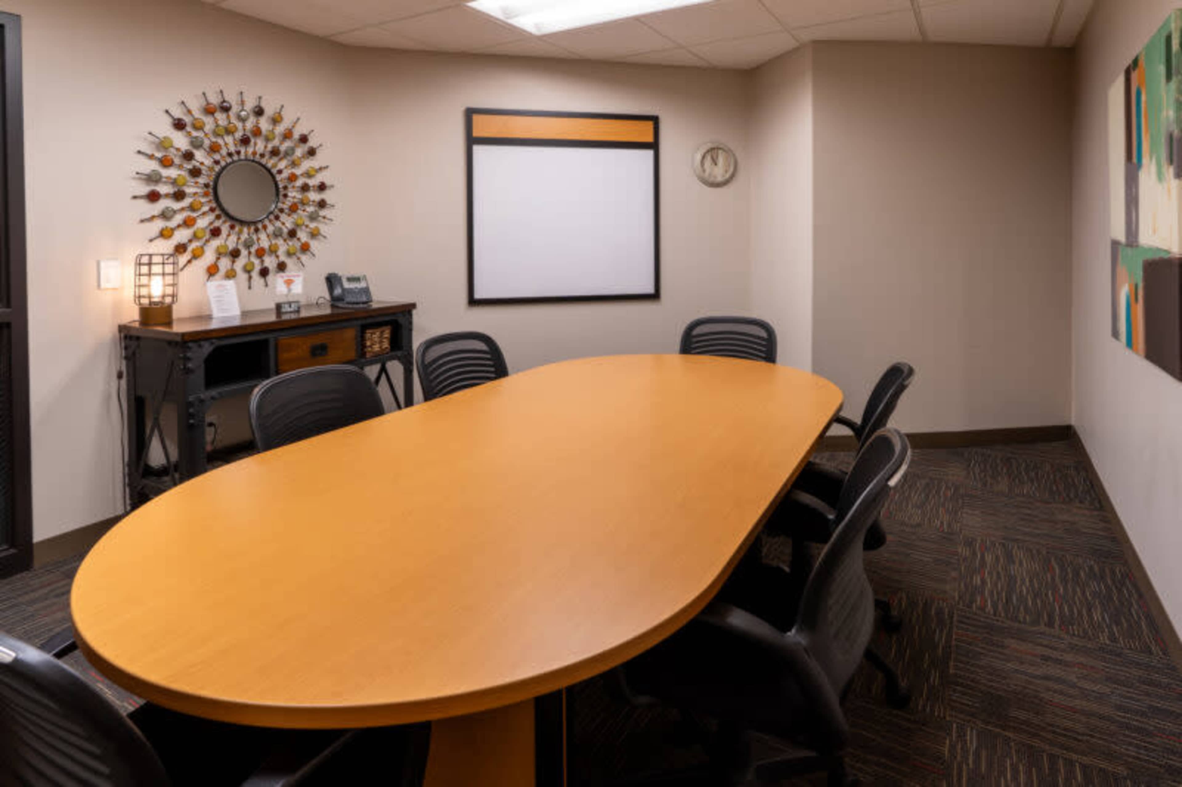 A conference room features an oval wooden table surrounded by black chairs, with a wall-mounted mirror and a blank whiteboard.