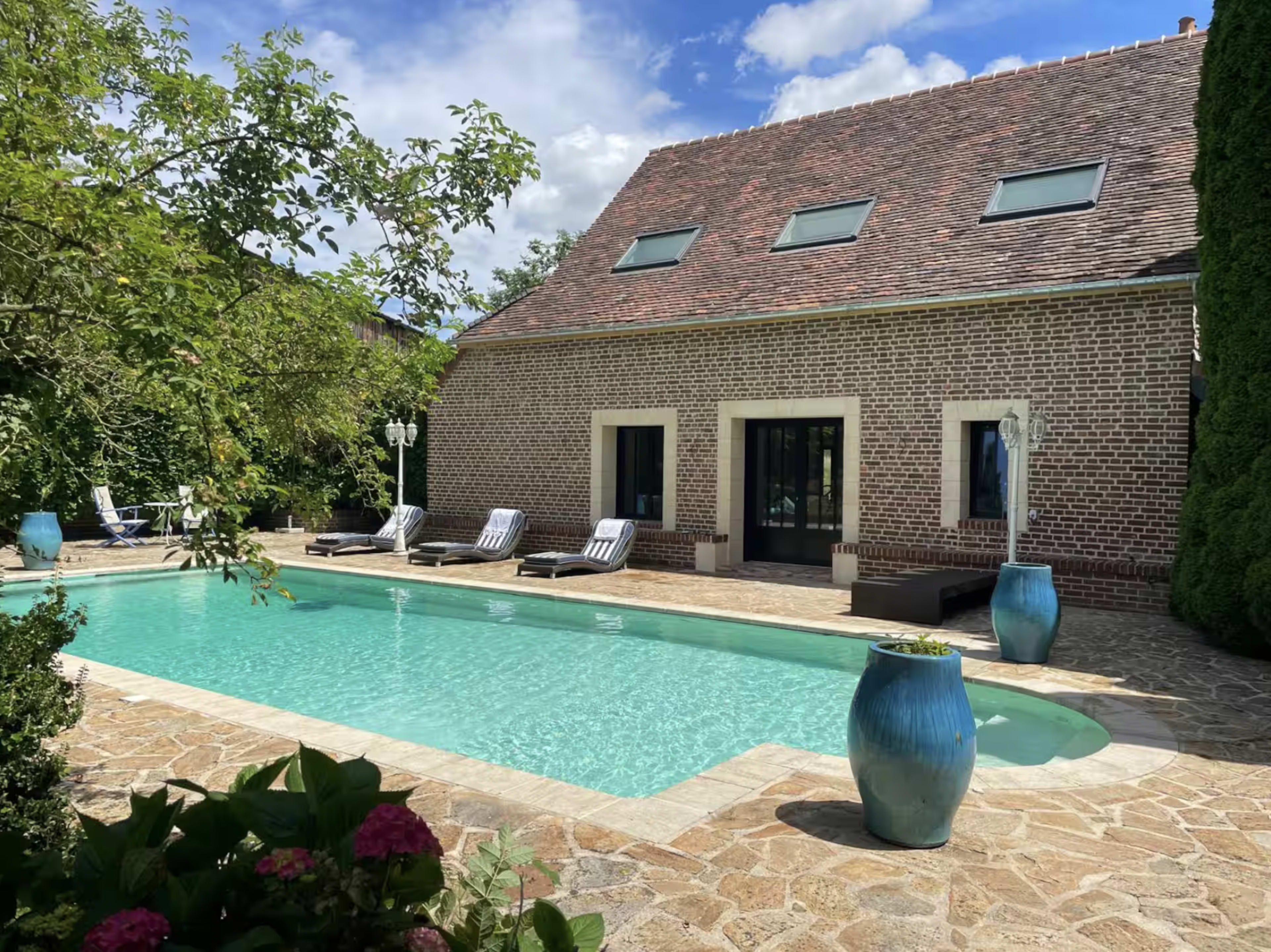 A modern house with a sloped roof sits beside a rectangular swimming pool, surrounded by lounge chairs and greenery.