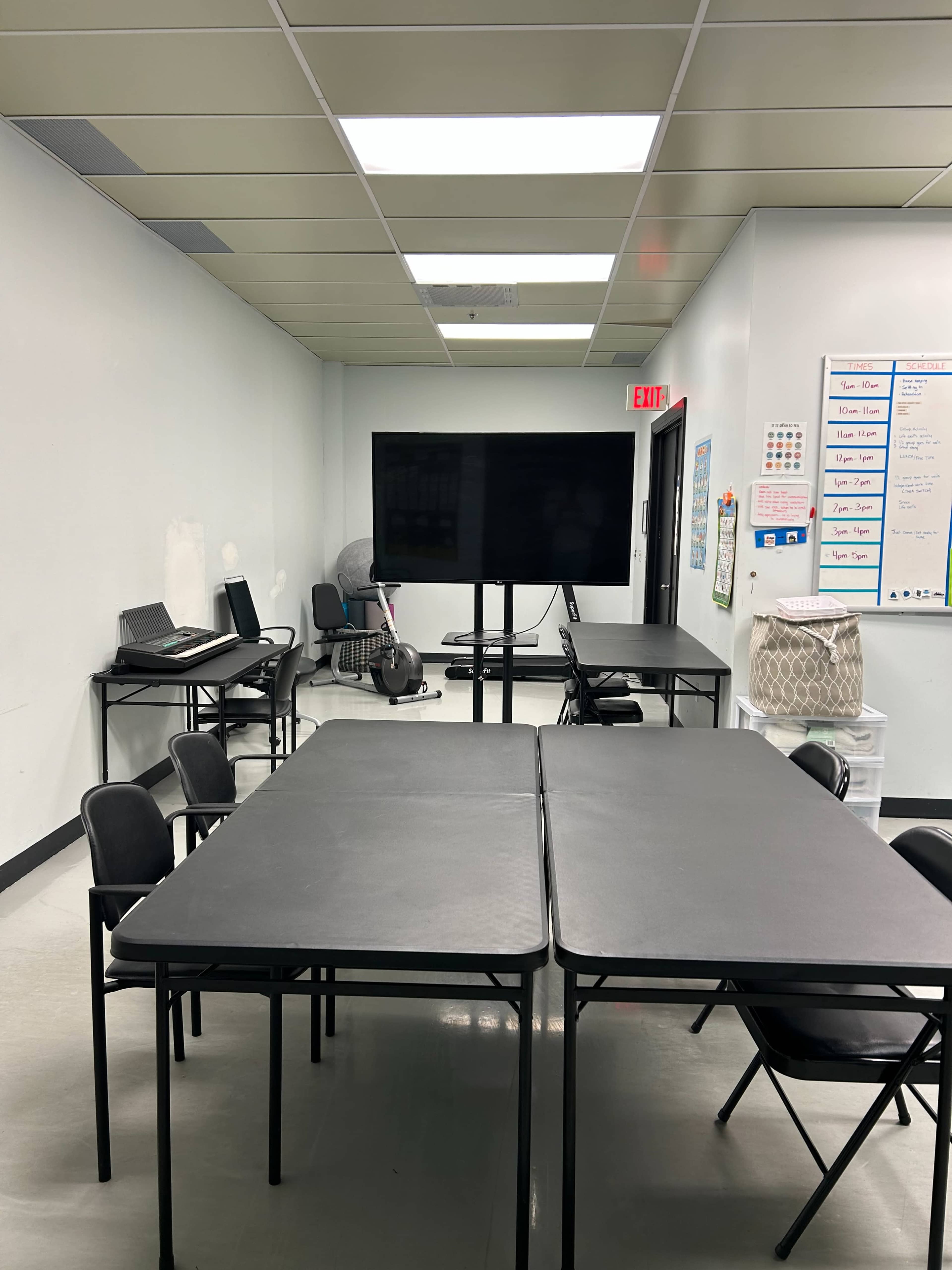 A classroom setting features several black tables arranged in the center, with a large screen and chairs positioned around the room.