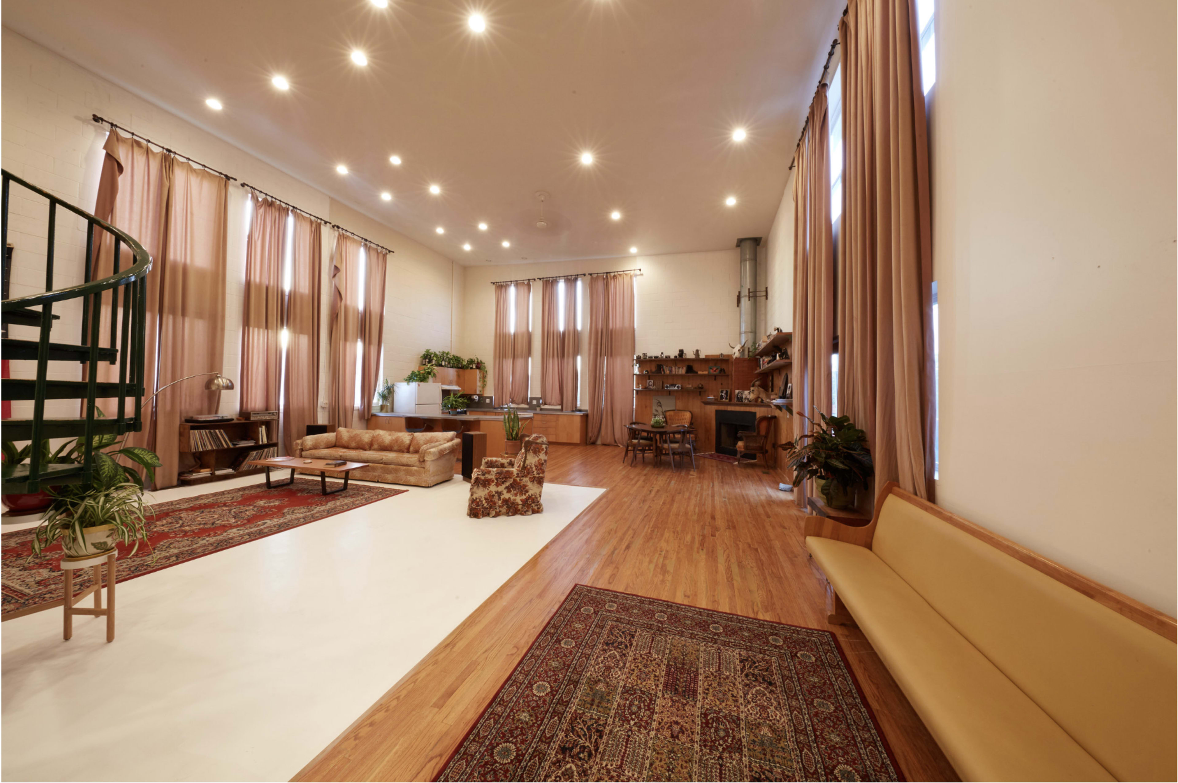A spacious, well-lit room with wooden flooring, large windows, and various furnishings including a sofa, a dining table, and plants.