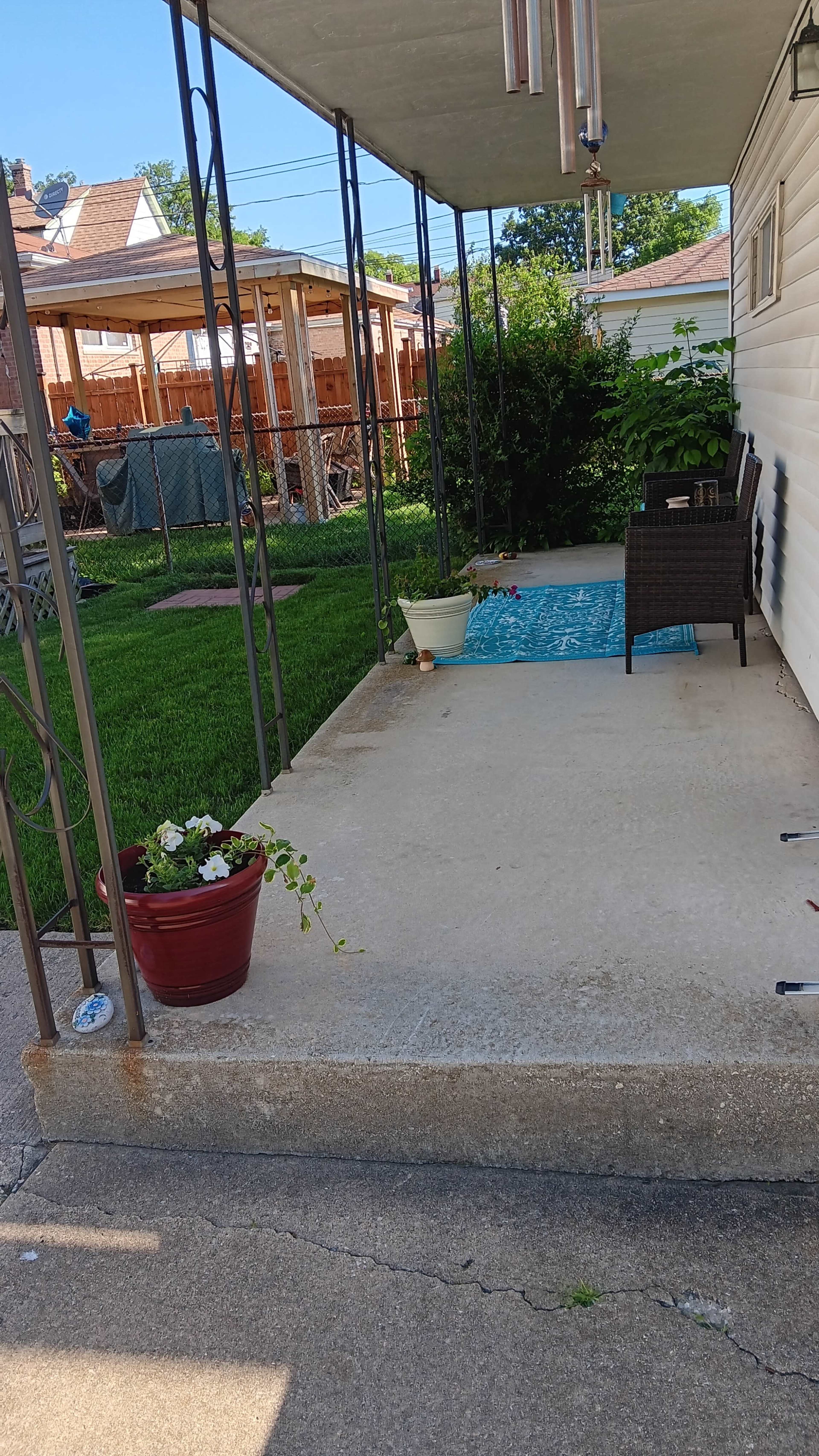 A concrete patio is surrounded by potted plants and leads to a grassy yard with a wooden structure in the background.