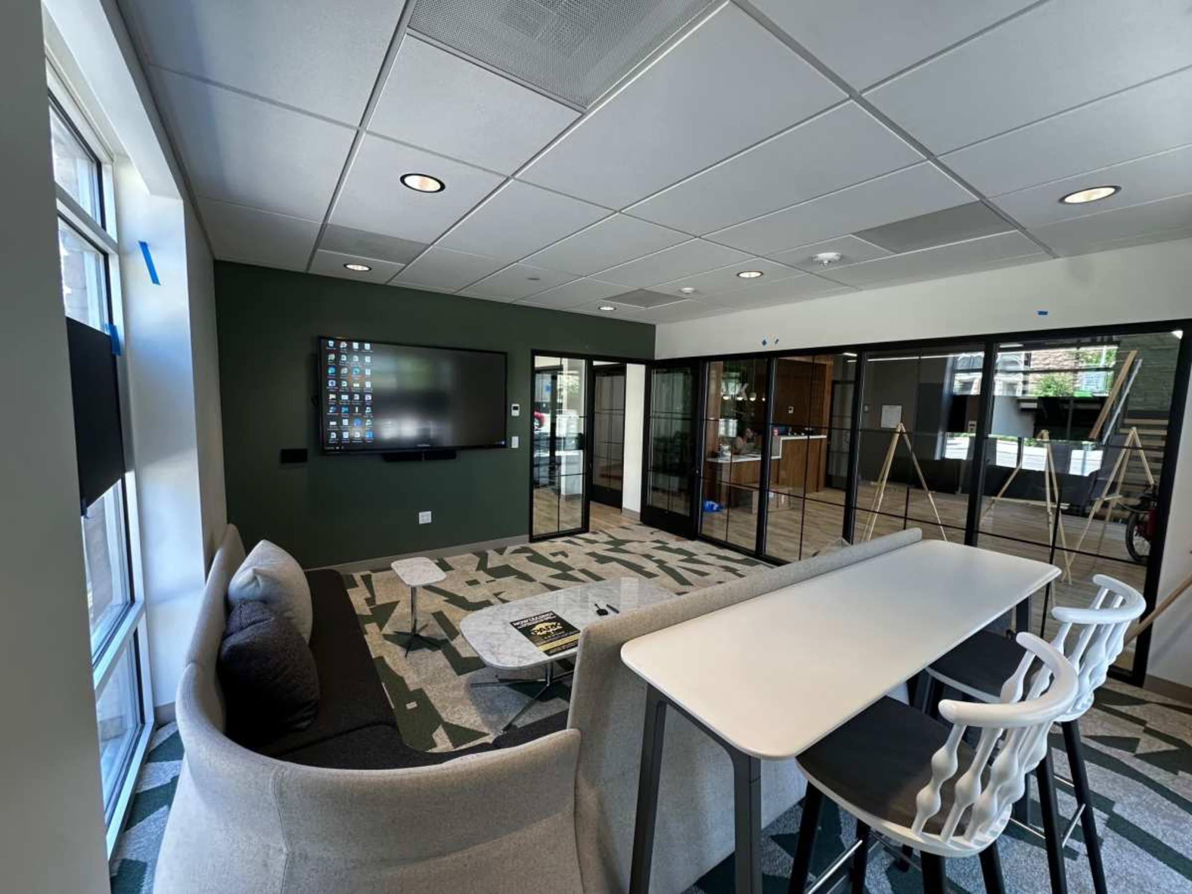 The image shows a modern office lounge area with a gray sofa, a white table, a large wall-mounted screen, and glass partitions leading to a conference room.