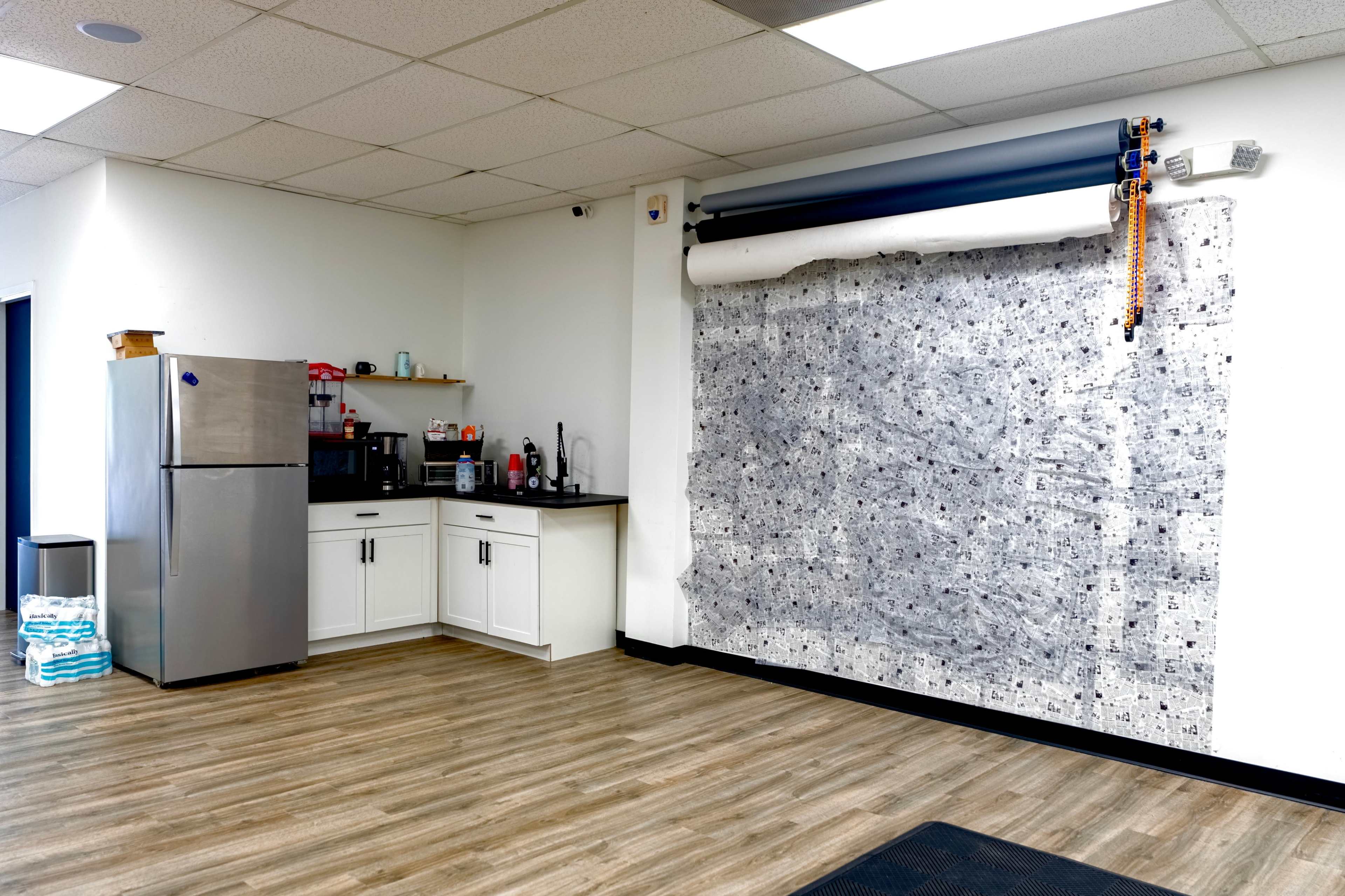 A kitchenette is located in one corner of a spacious room, featuring a refrigerator, microwave, and a large roll of patterned fabric mounted on the wall.