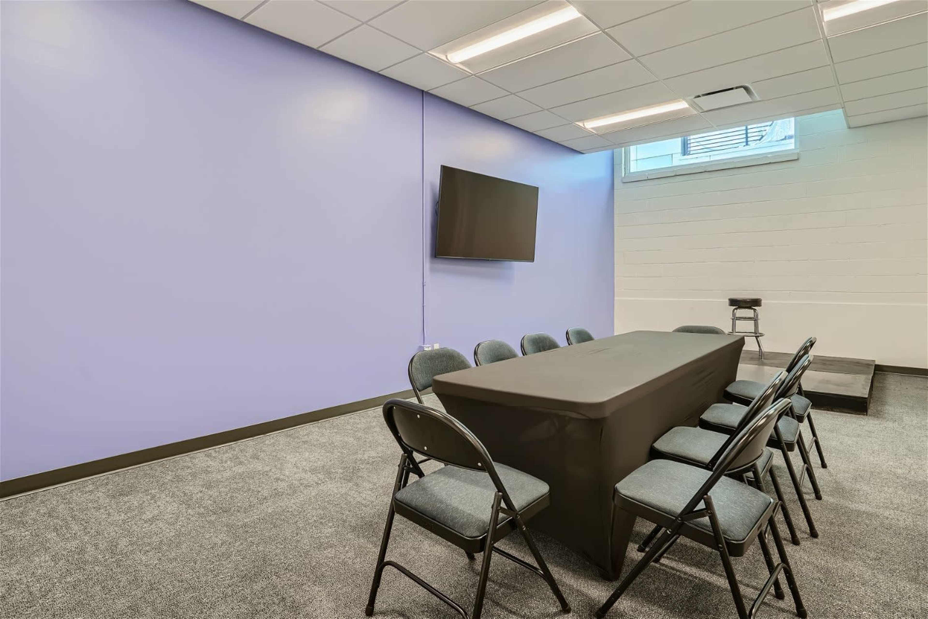 A conference room with a long black table surrounded by eight chairs, a television mounted on the wall, and a light purple wall color.