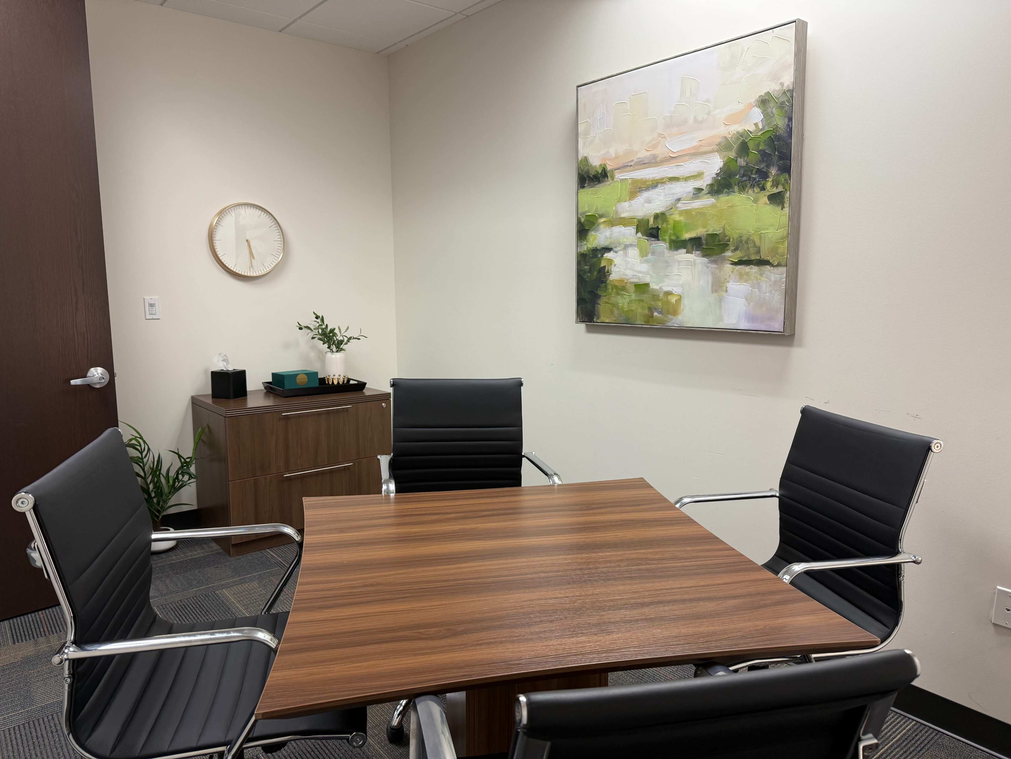 The image shows a small meeting room featuring a square wooden table surrounded by four black chairs, with a painting of a landscape on the wall.
