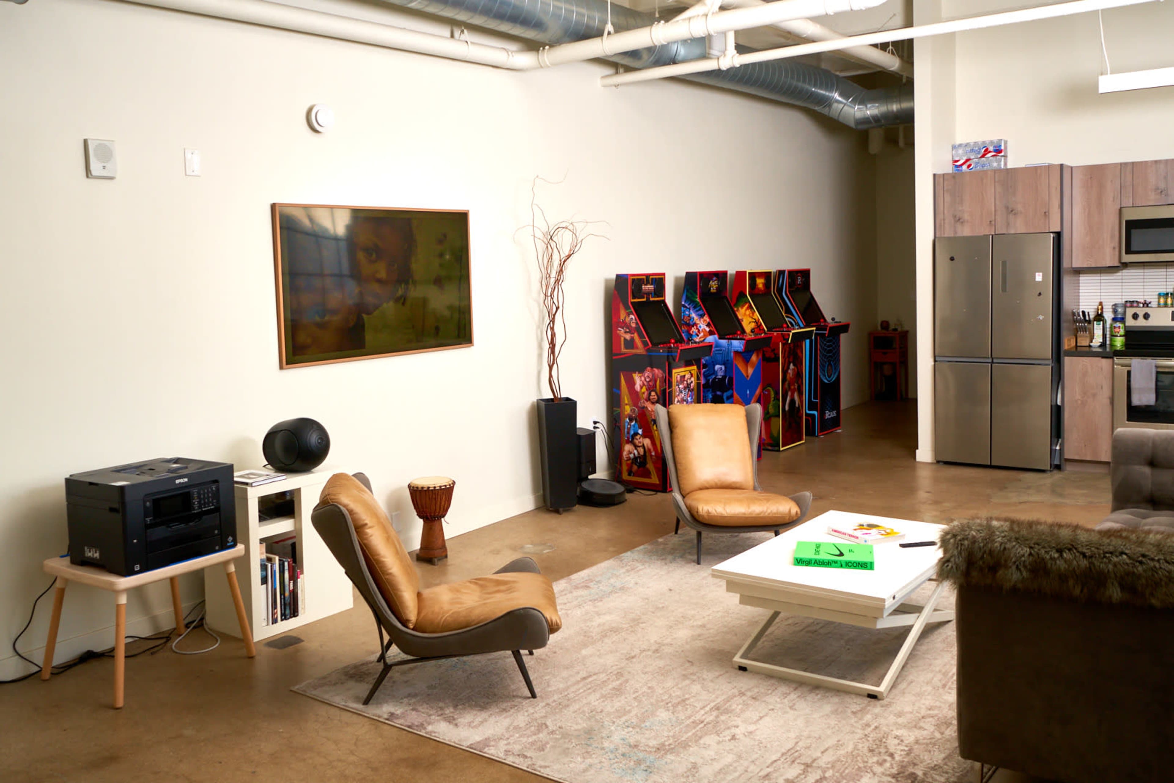 A spacious living area featuring two leather chairs, a large coffee table, and a kitchenette with modern appliances, alongside vintage arcade game machines in the background.