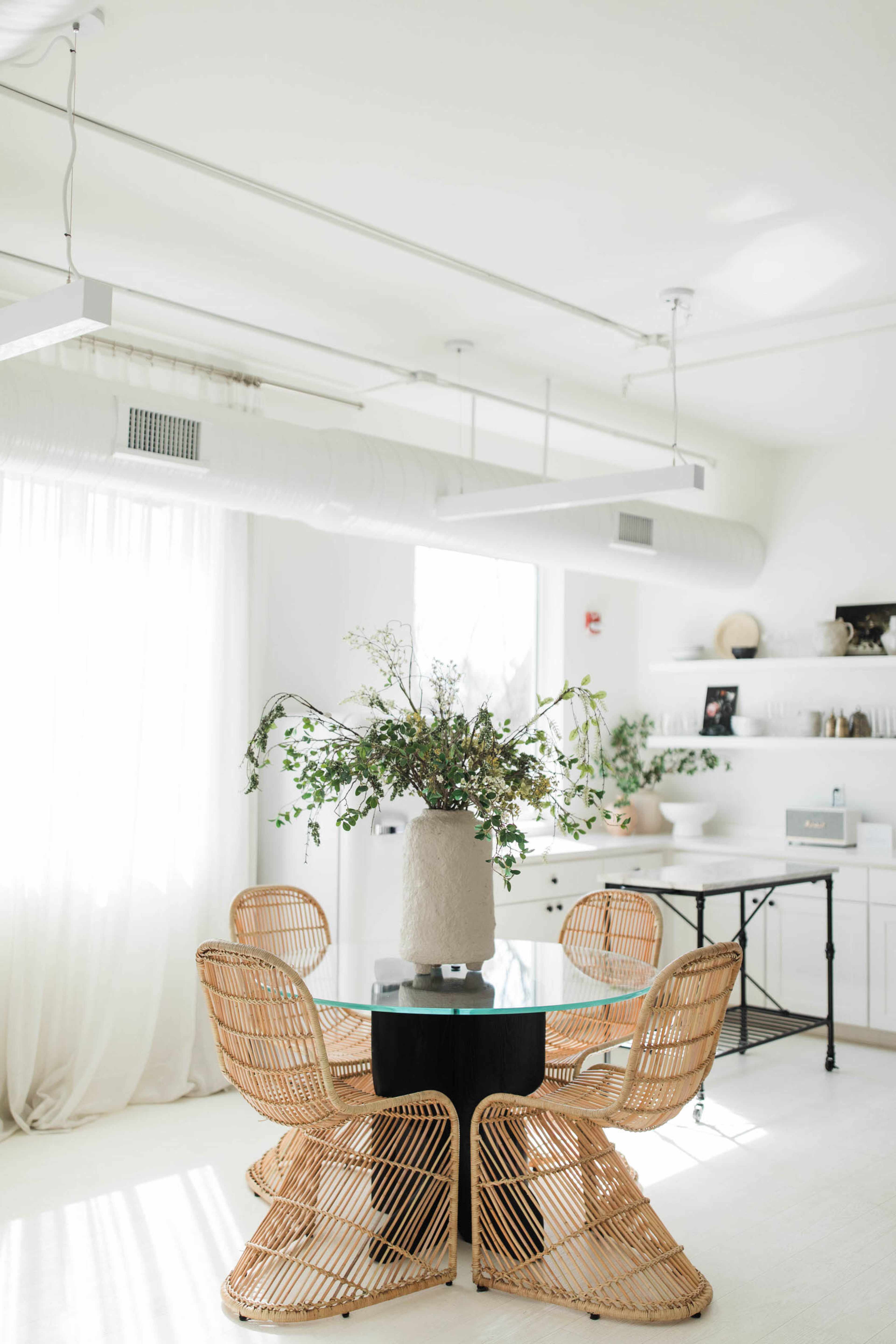 A dining area features a round glass table surrounded by rattan chairs, with a large vase of greenery as the centerpiece.