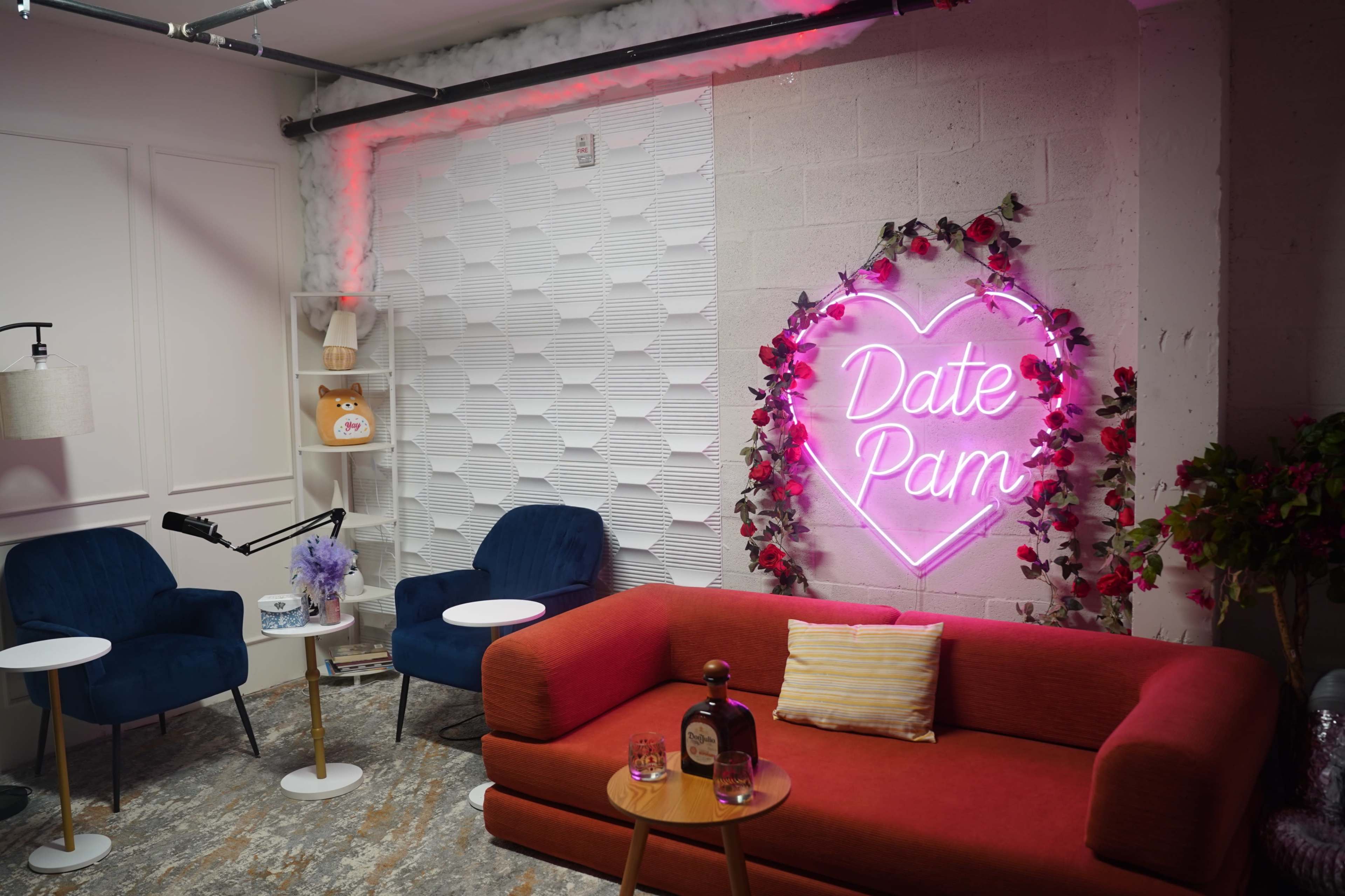 The image shows a cozy interior space featuring a red sofa, two blue chairs, small white tables, and a neon sign that reads "Date Pam" adorned with flowers.