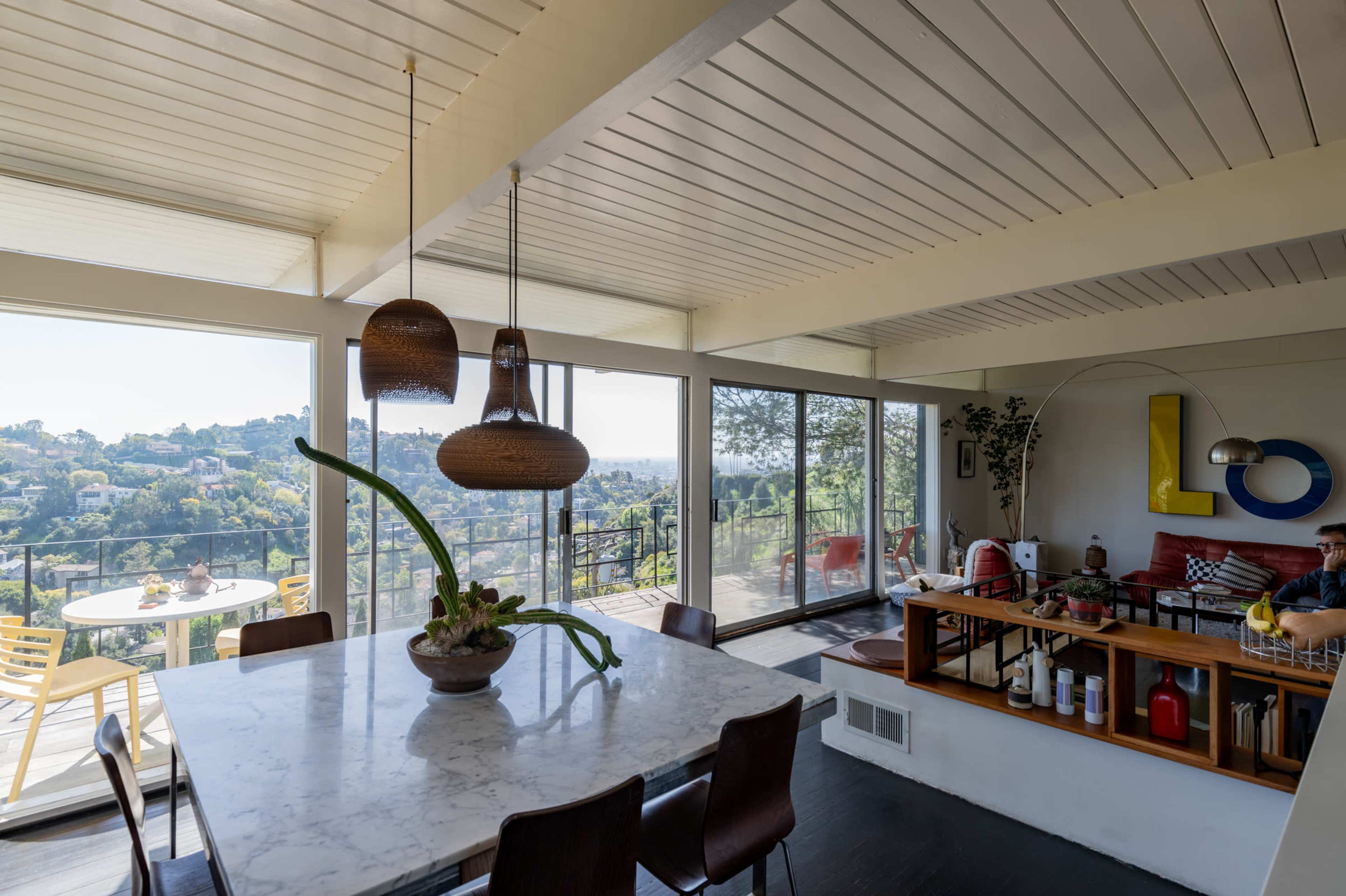 A modern dining area features a marble table, wooden chairs, and large windows offering a view of the landscape beyond.