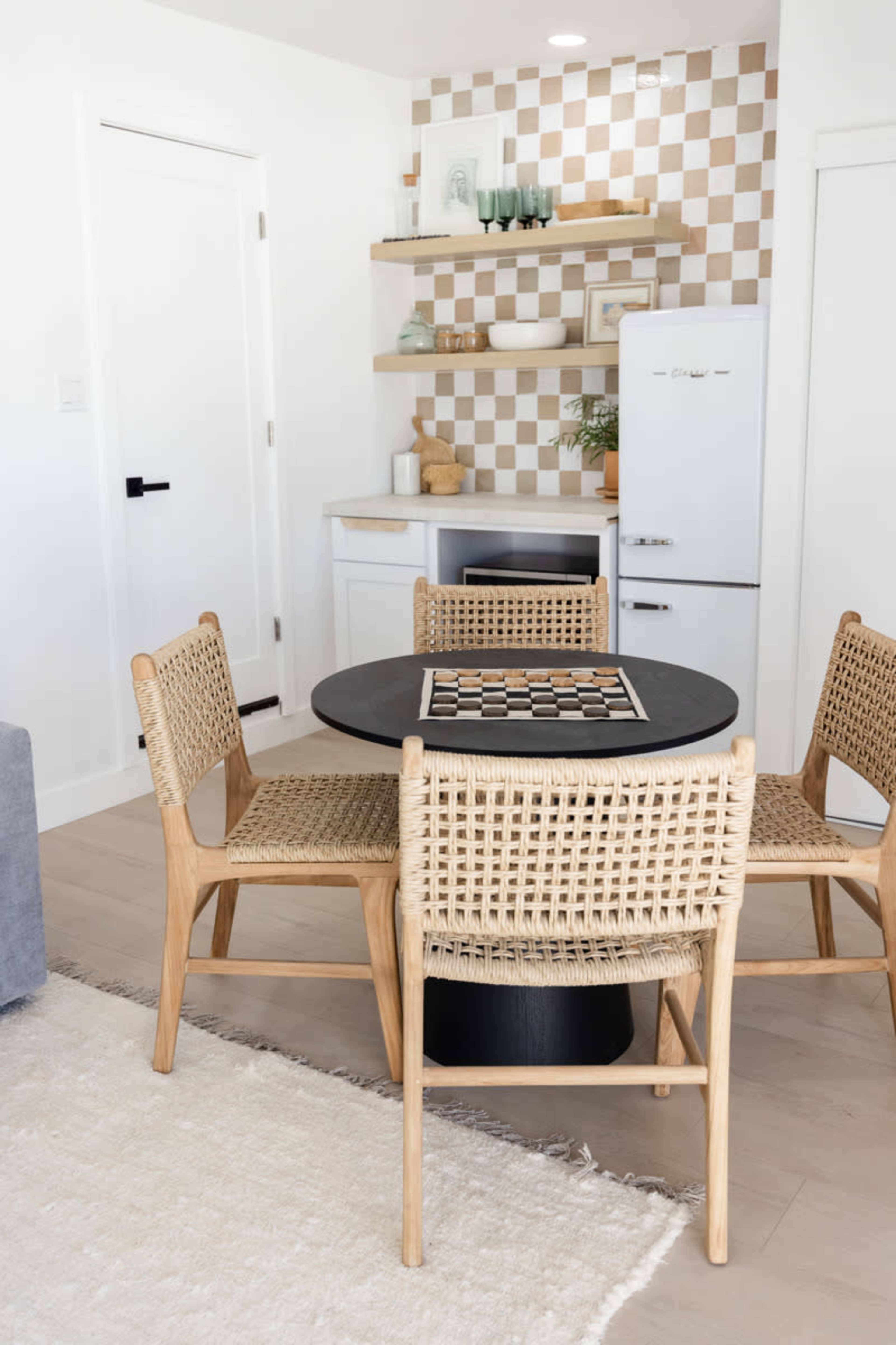 A dining area with a round black table set with a chessboard, surrounded by four rattan chairs, against a backdrop of a minimal kitchen with checkered wall tiles.