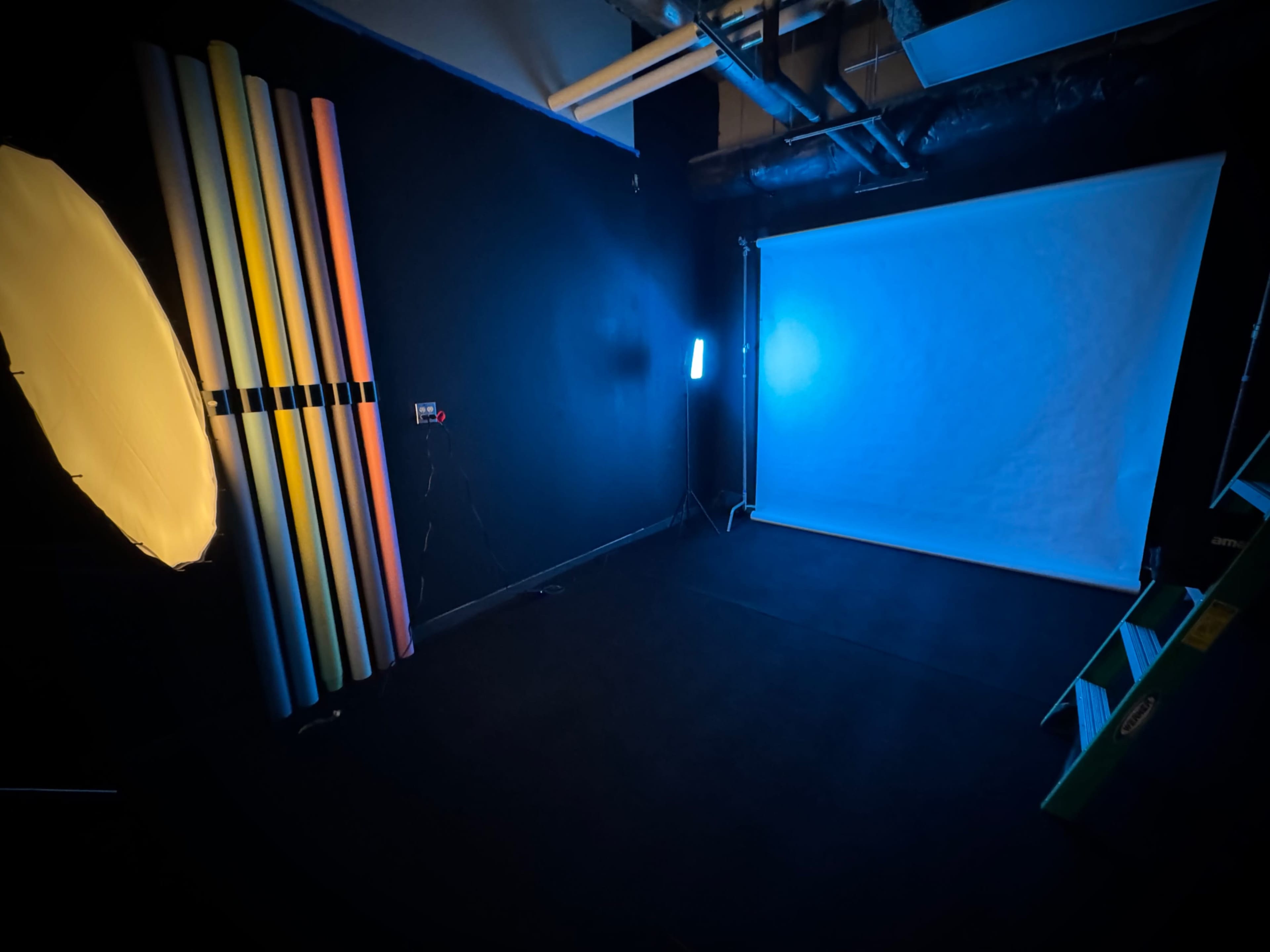 The image shows a photo studio setup featuring a white backdrop, various colored paper rolls leaning against a black wall, and lighting equipment in a dimly lit environment.