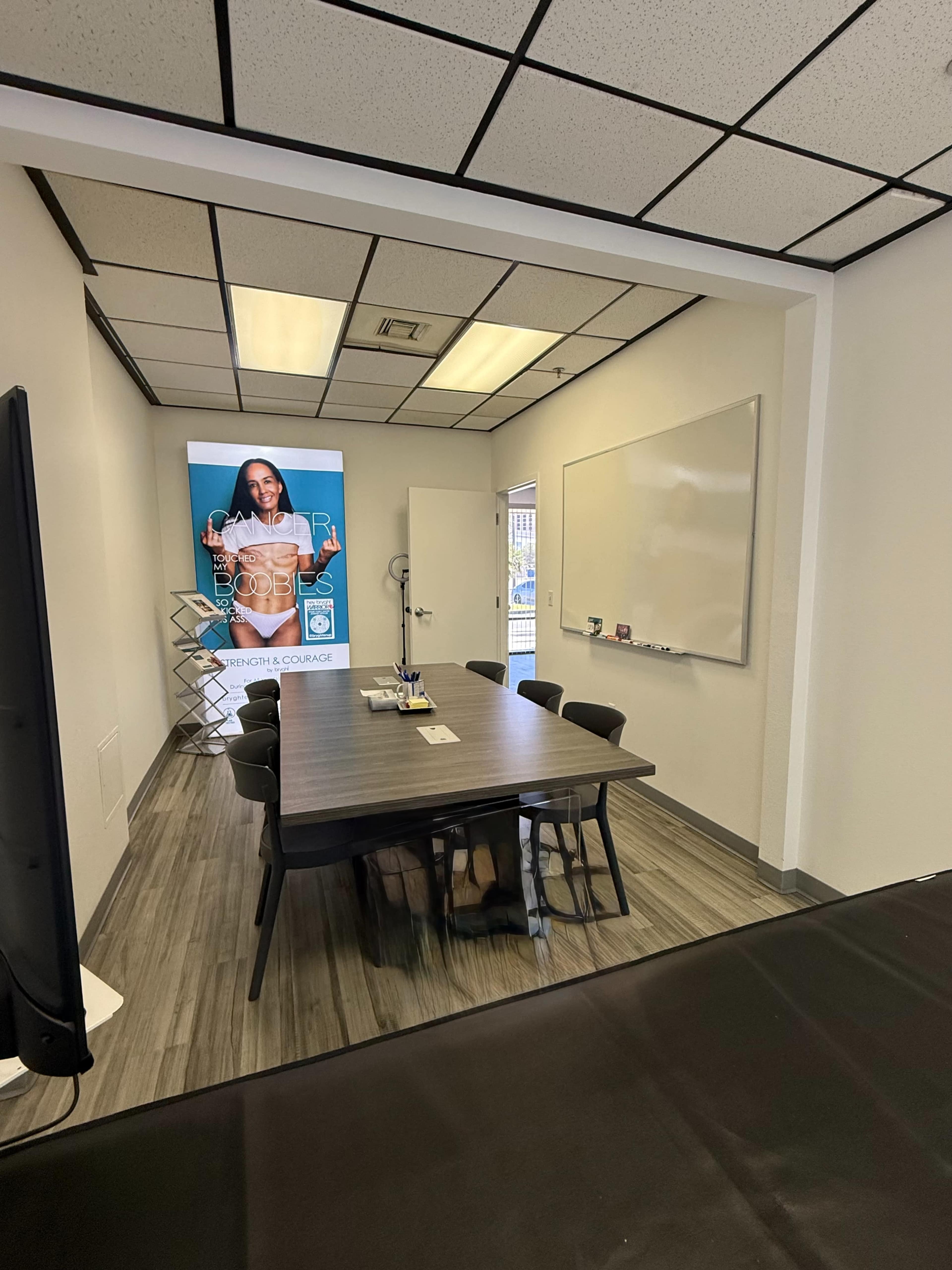 The image shows a conference room with a large table surrounded by chairs, a whiteboard on one wall, and a poster featuring a person on another wall.