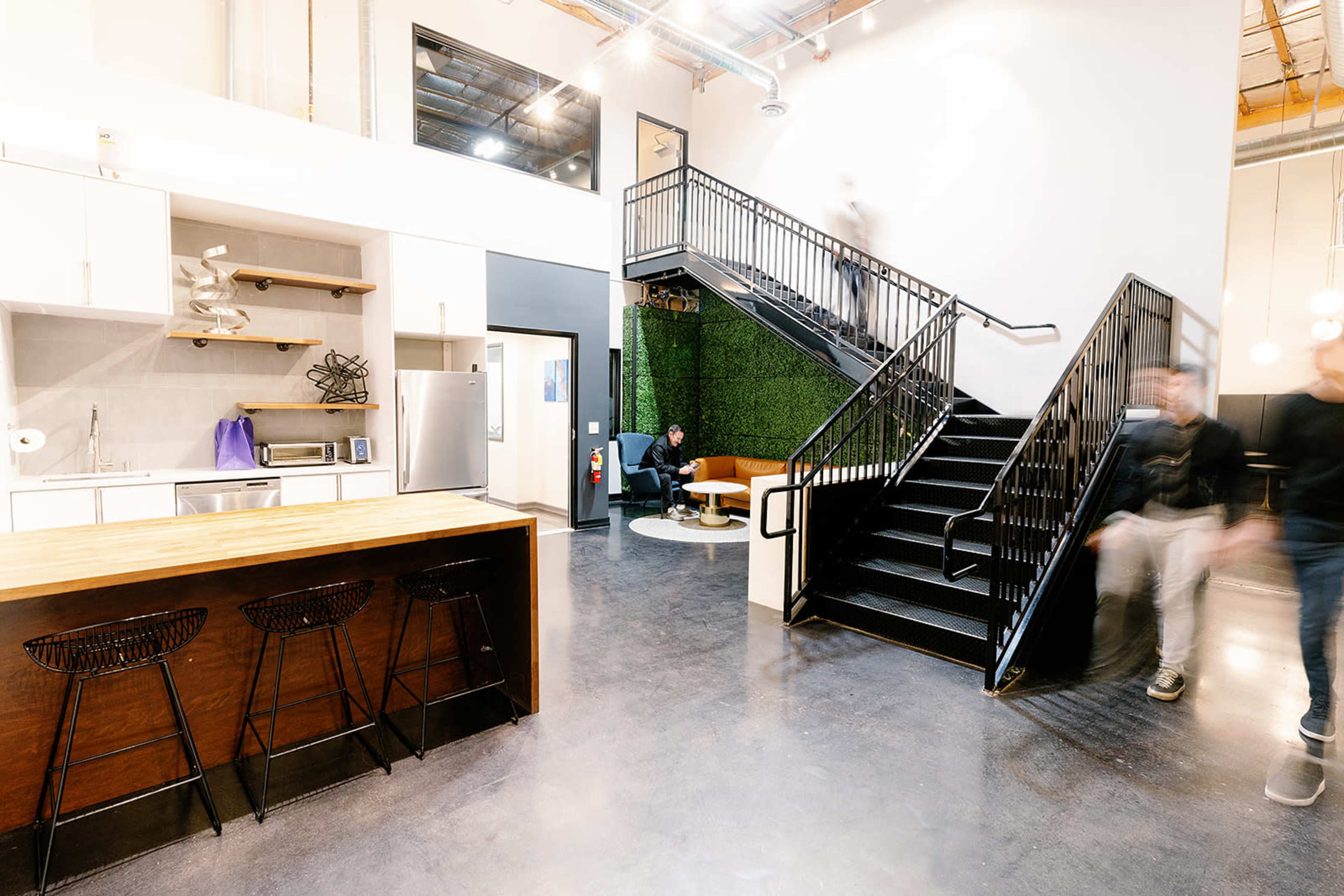 A modern open-plan workspace featuring a kitchen area with bar stools, a green wall, and a staircase leading to an upper level.