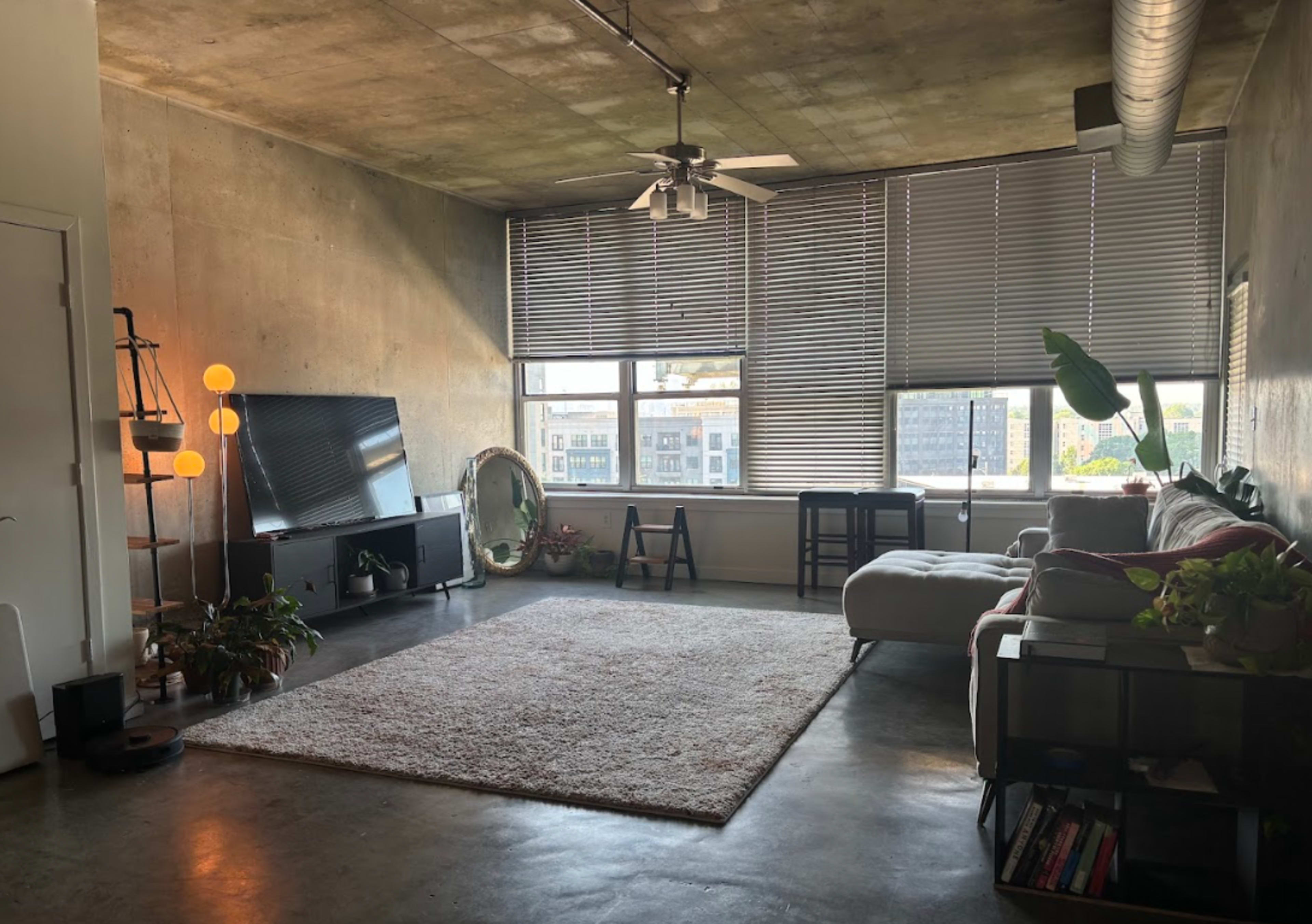 The image shows a spacious, modern living room with large windows, a light-colored sofa, a television, and various plants arranged throughout the space.