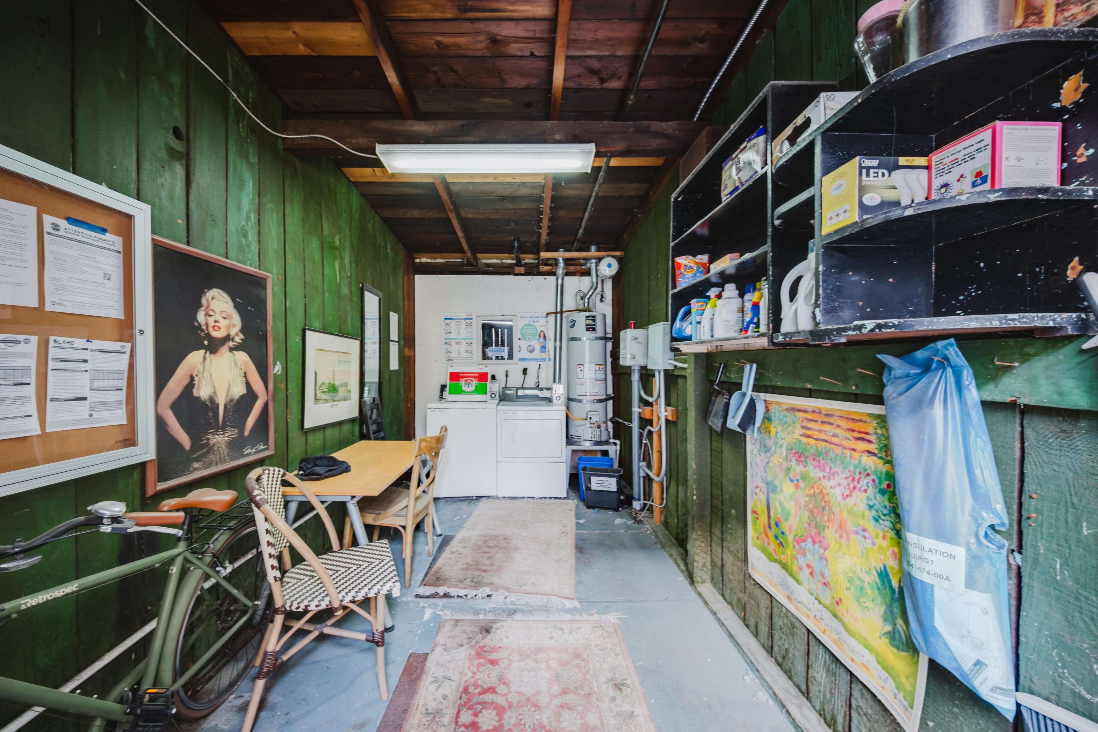 The image shows a narrow room with green wooden walls, featuring a dining table, a bicycle, laundry appliances, and various shelves stocked with cleaning supplies.