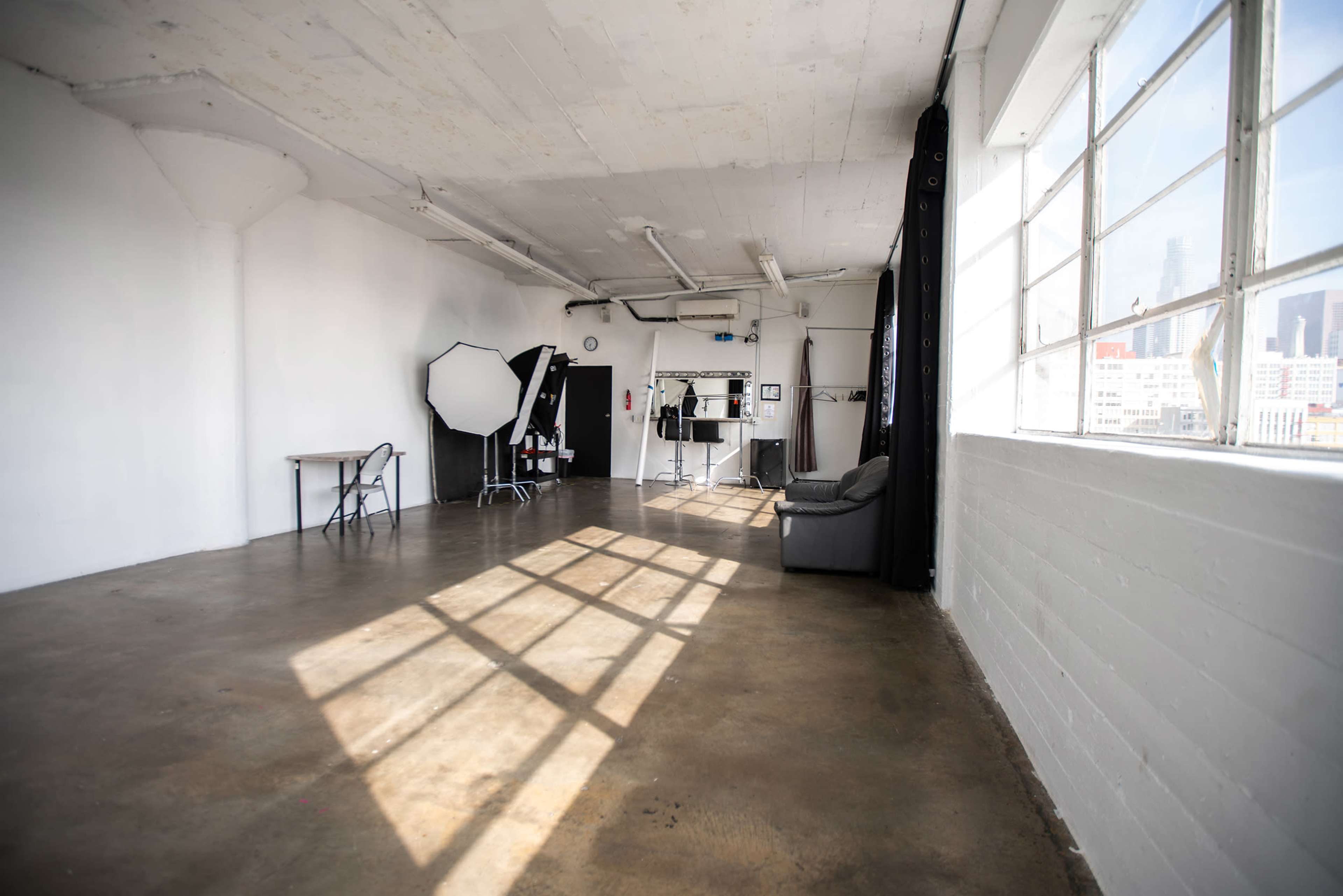 A spacious, empty studio with large windows, a simple table, a folding chair, and lighting equipment.