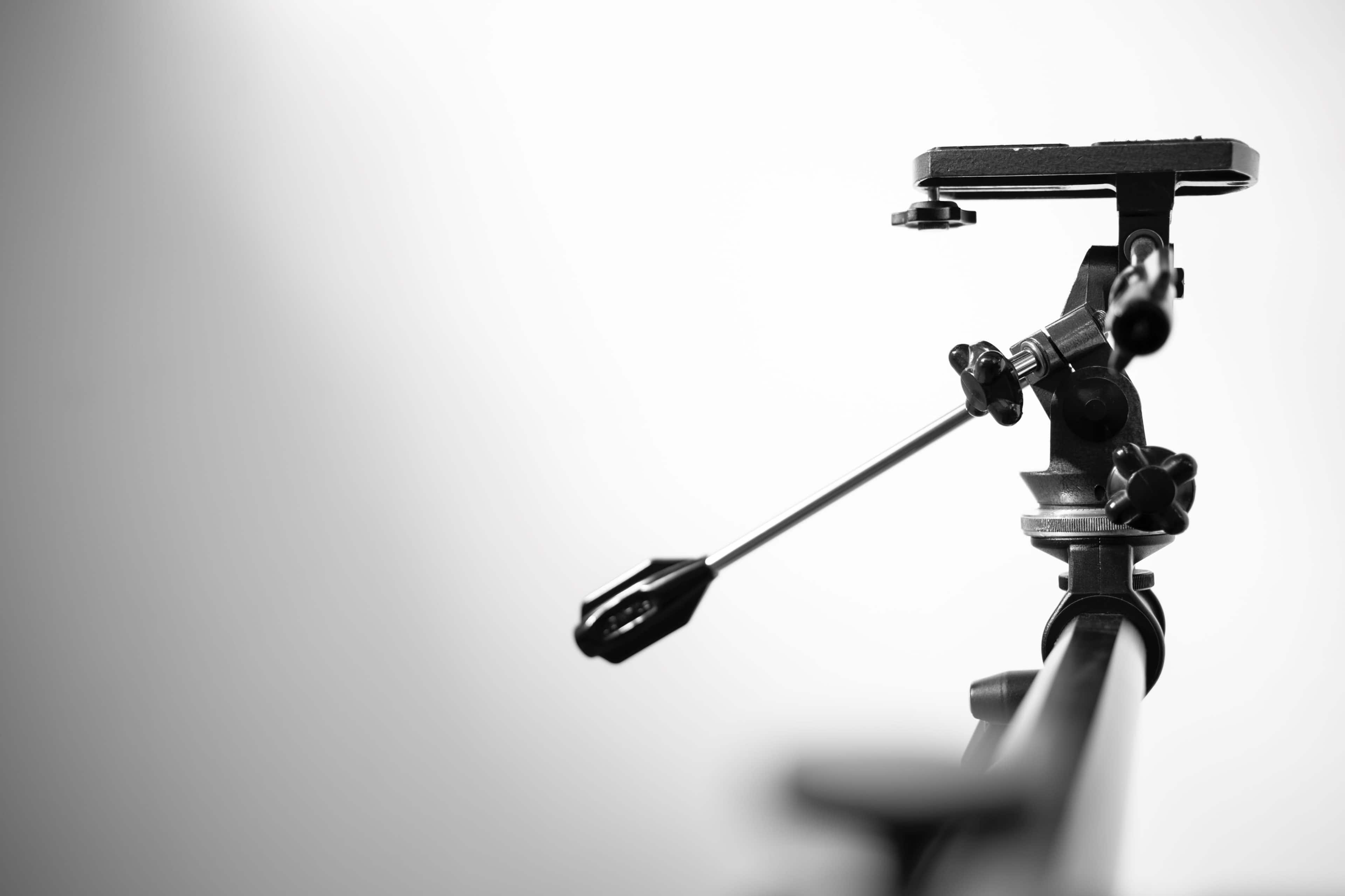 A close-up of a camera tripod head with an adjustable arm and quick-release plate against a gradient background.