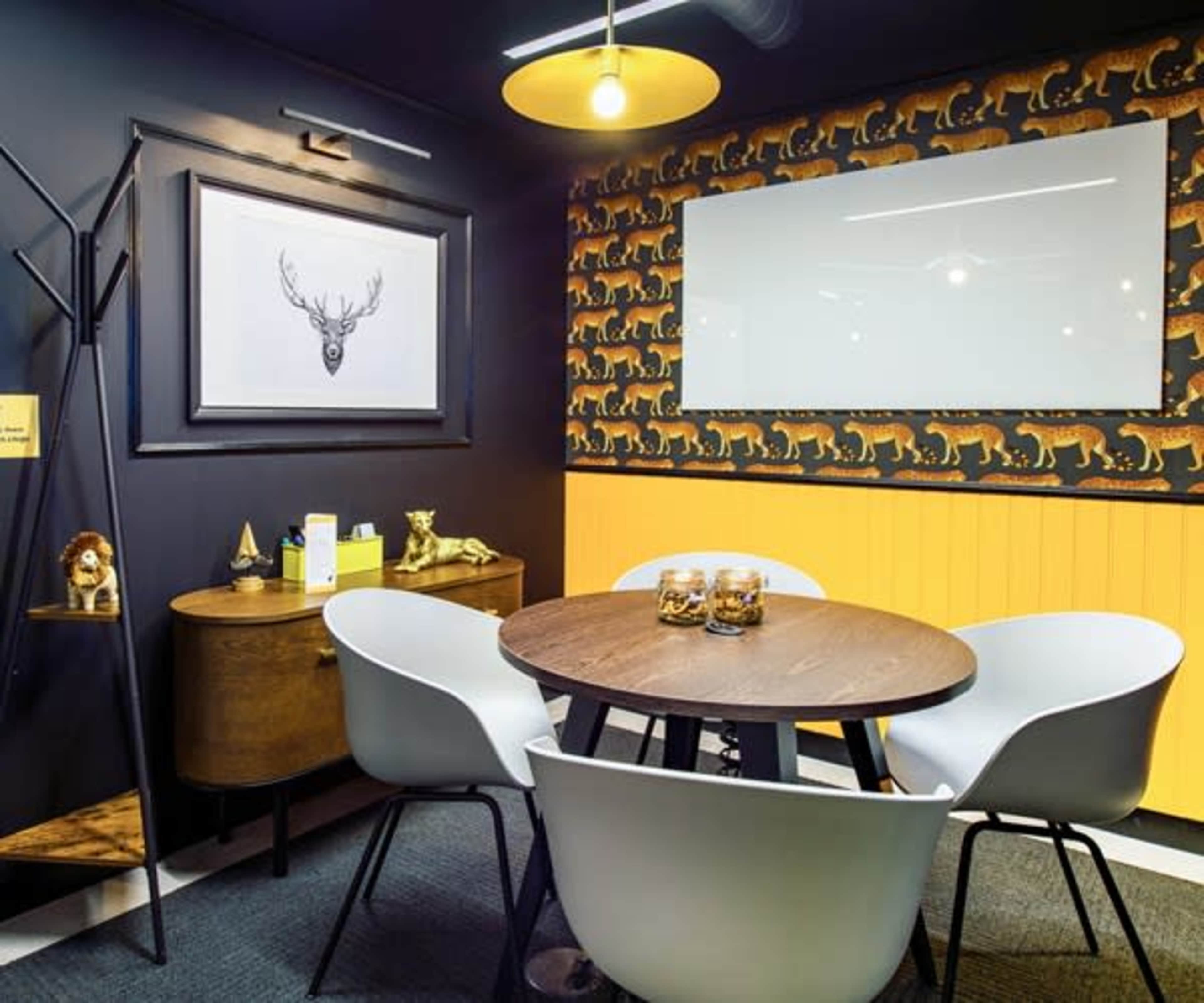 A small meeting room features a round wooden table surrounded by white chairs, a deer print on the wall, and a patterned wallpaper depicting animals.