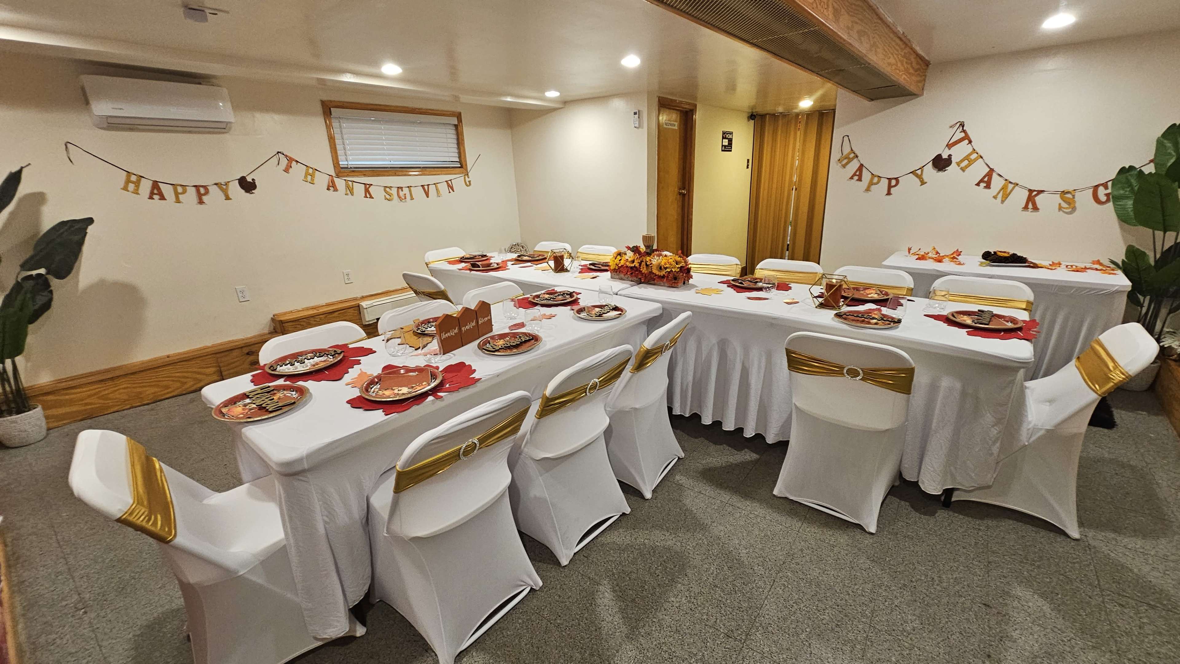 A dining area is set up with tables adorned in white tablecloths and gold accents, featuring plates and decorations for a Thanksgiving celebration.