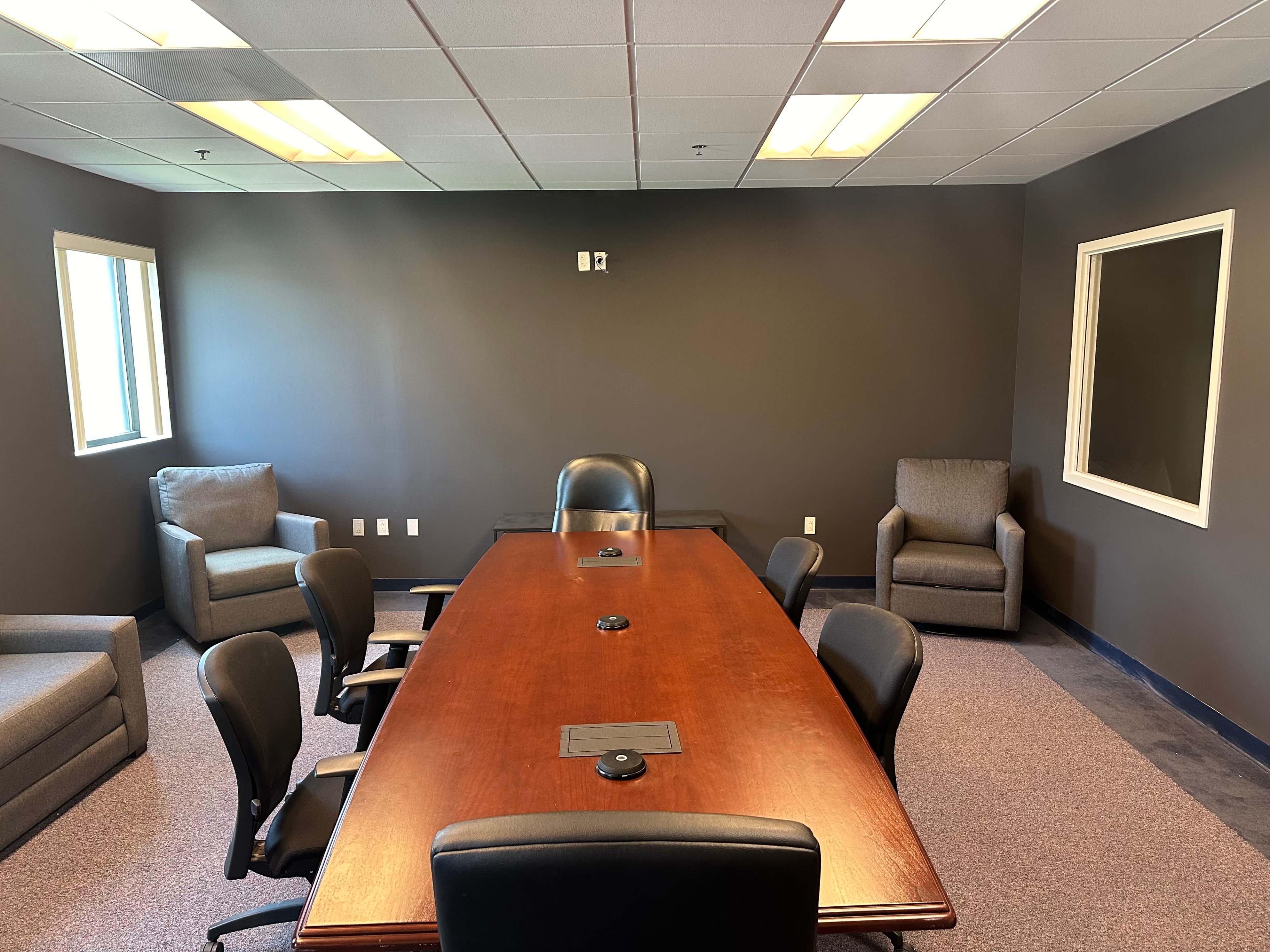 A meeting room with a large wooden table surrounded by black office chairs, flanked by two armchairs and a window.