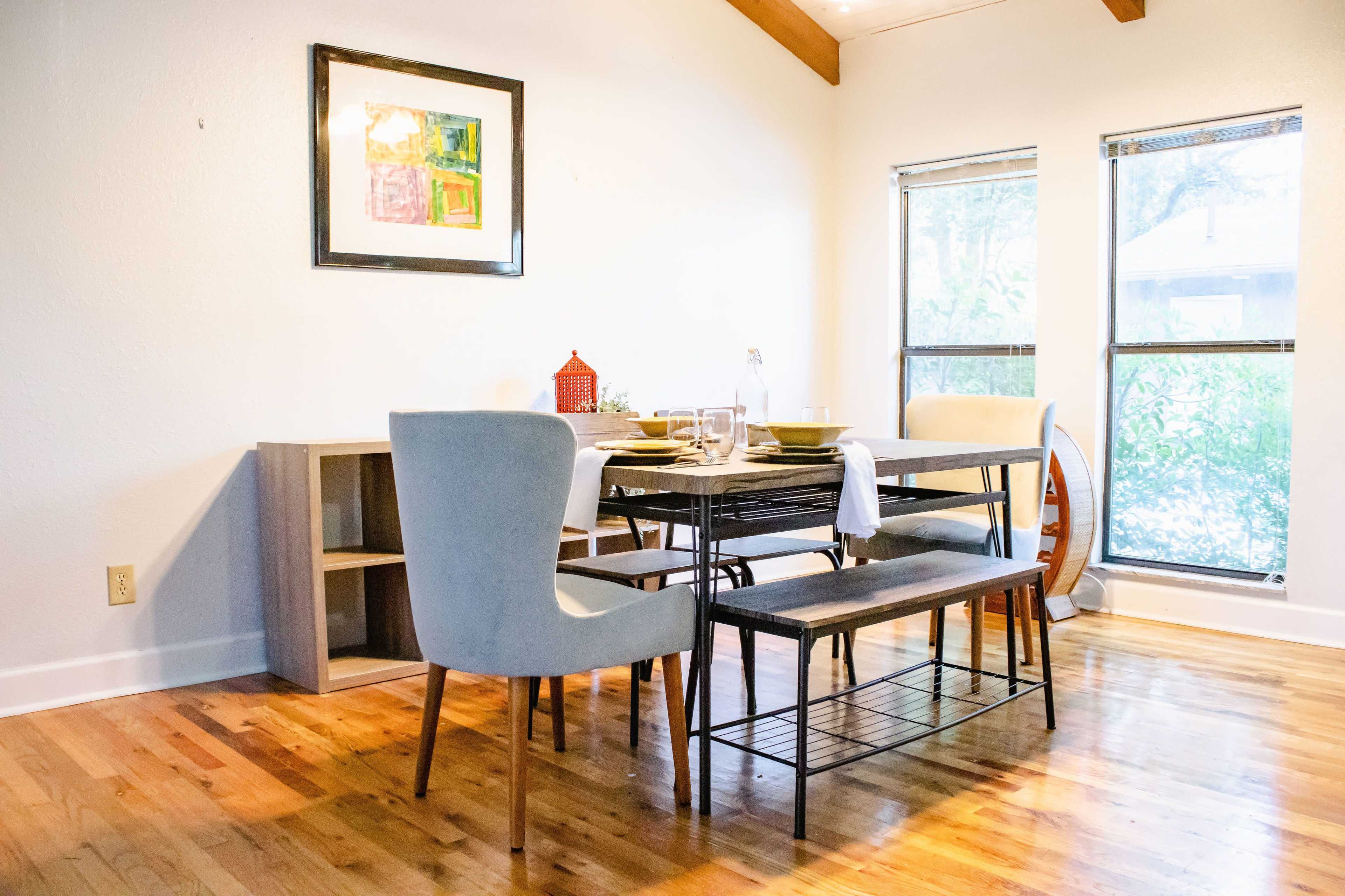A dining room features a table set with plates and glasses, accompanied by two chairs and a bench, with a sideboard and a window showing greenery outside.
