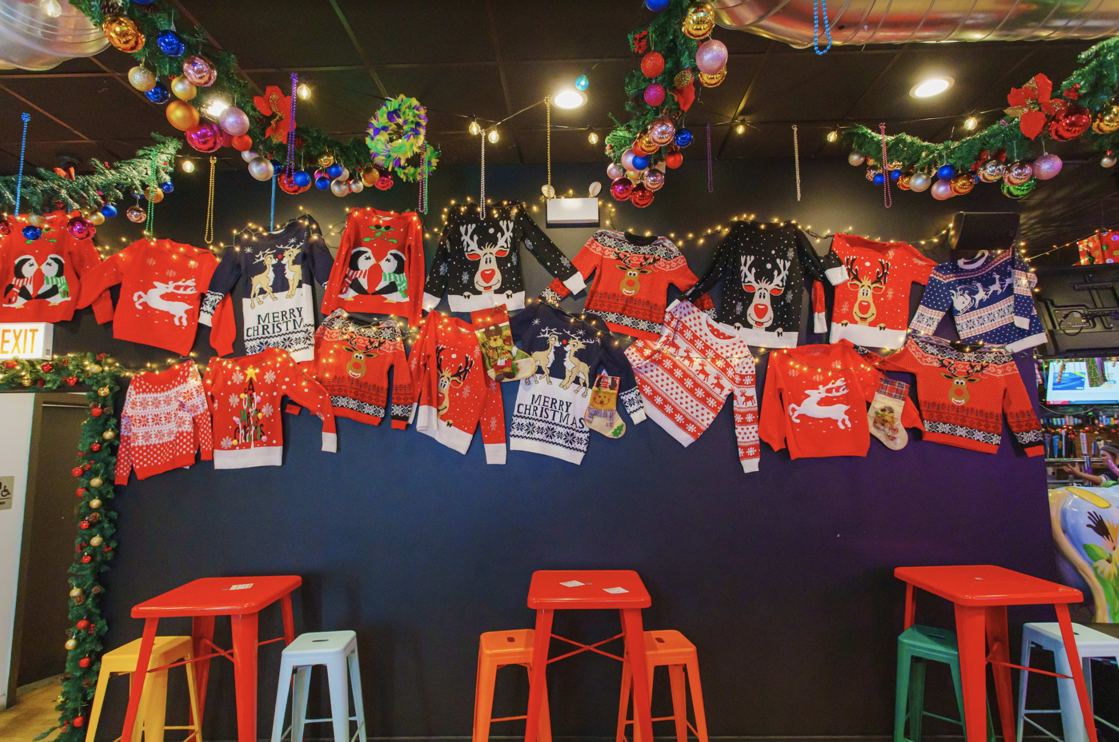 A wall decorated with colorful holiday-themed sweaters and Christmas ornaments, accompanied by red and green tables below.