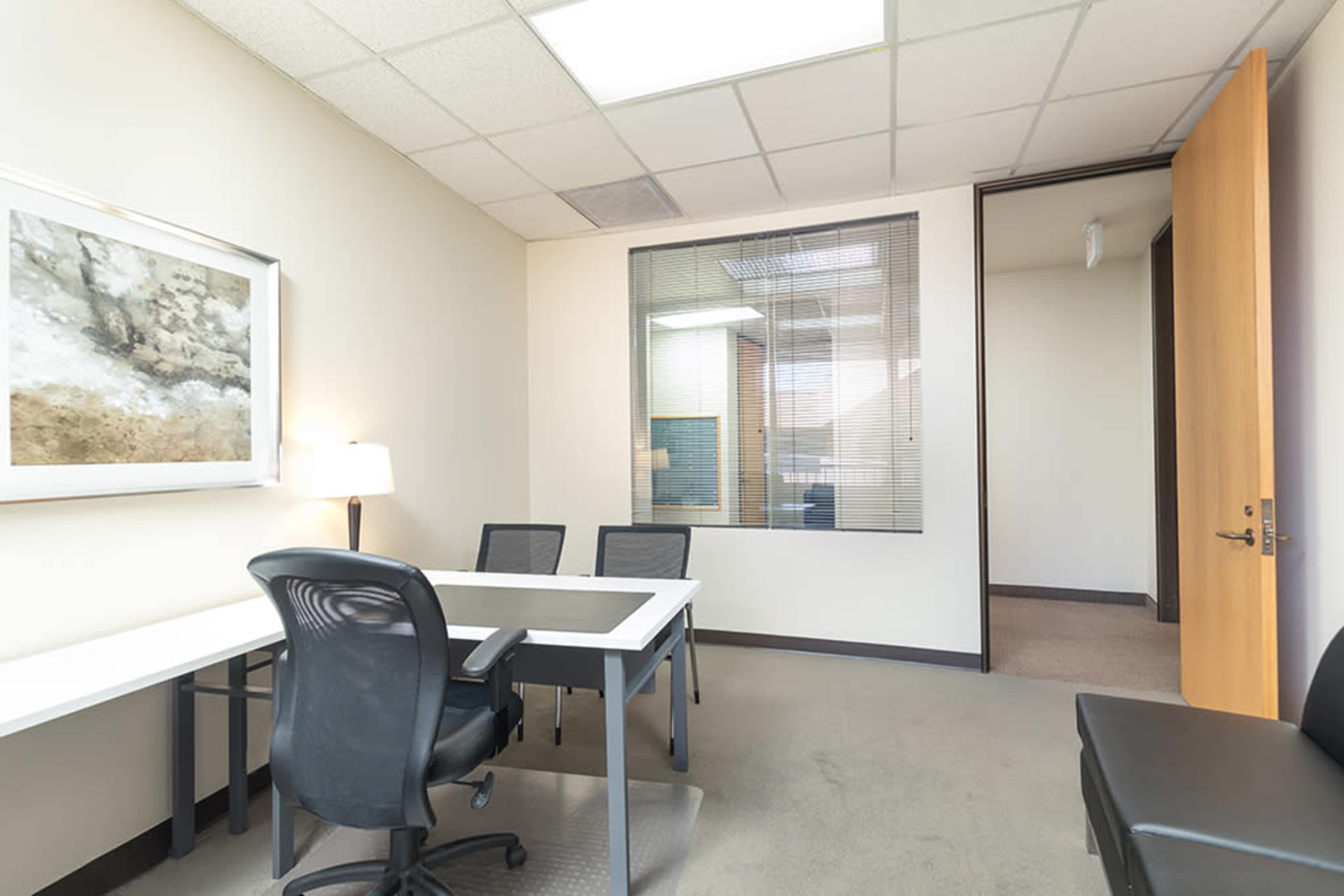 A small office room featuring a desk, three chairs, a lamp, and a glass-walled meeting area.