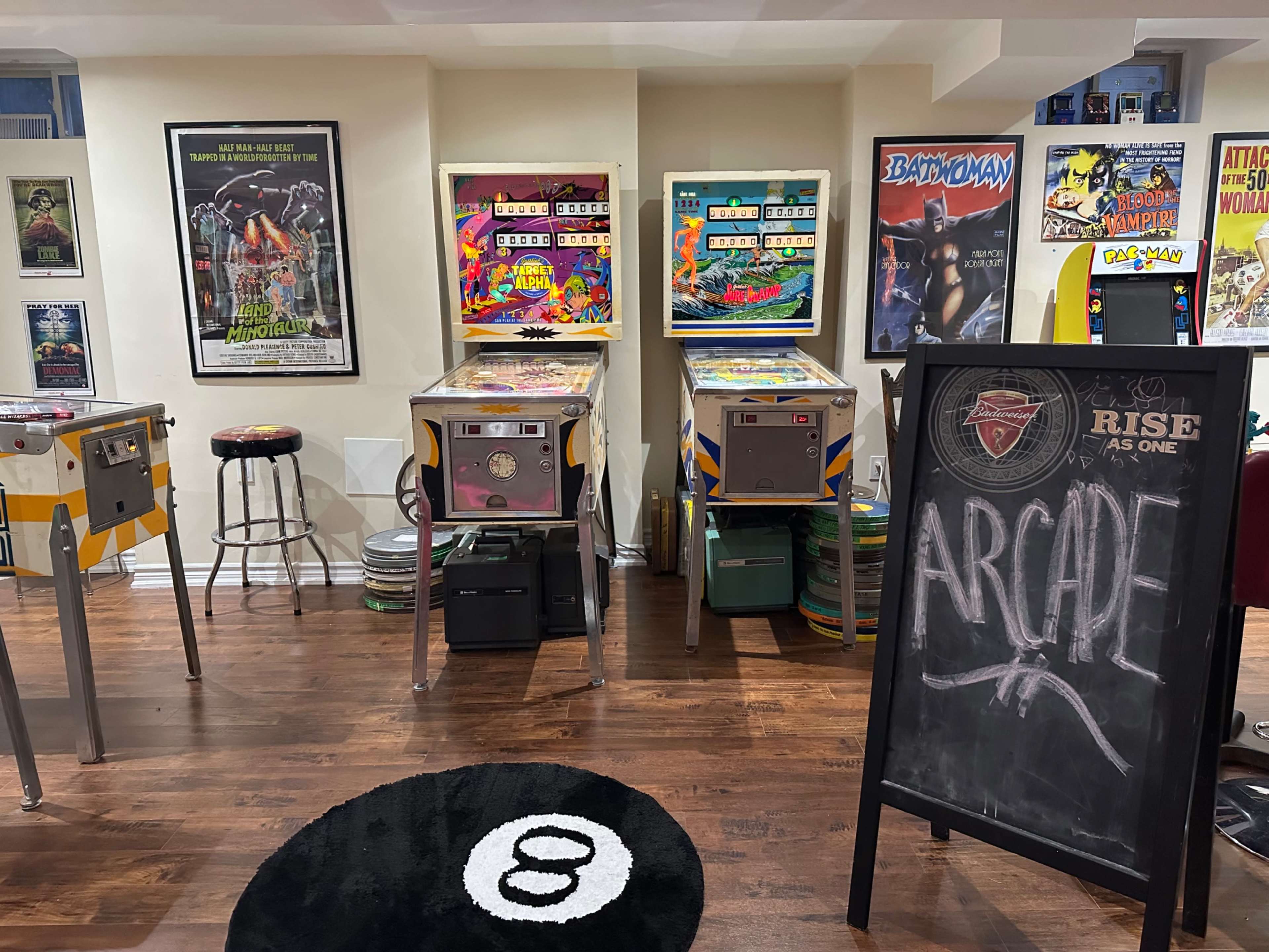 The image depicts a home arcade featuring several pinball machines, retro movie posters on the wall, and a black chalkboard sign that reads "ARCade."