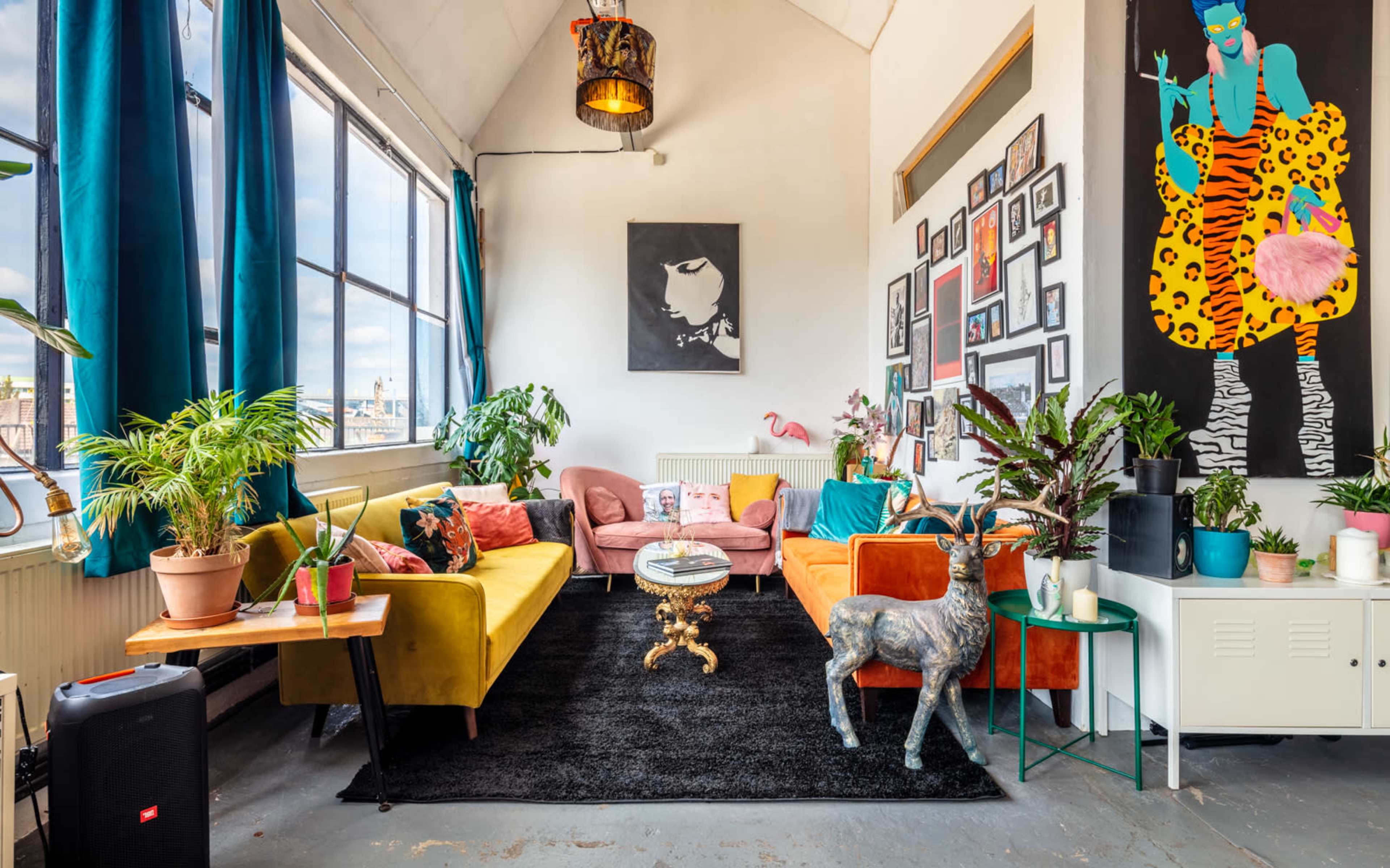A brightly lit living room with colorful sofas, various plants, and a large wall adorned with framed artworks.