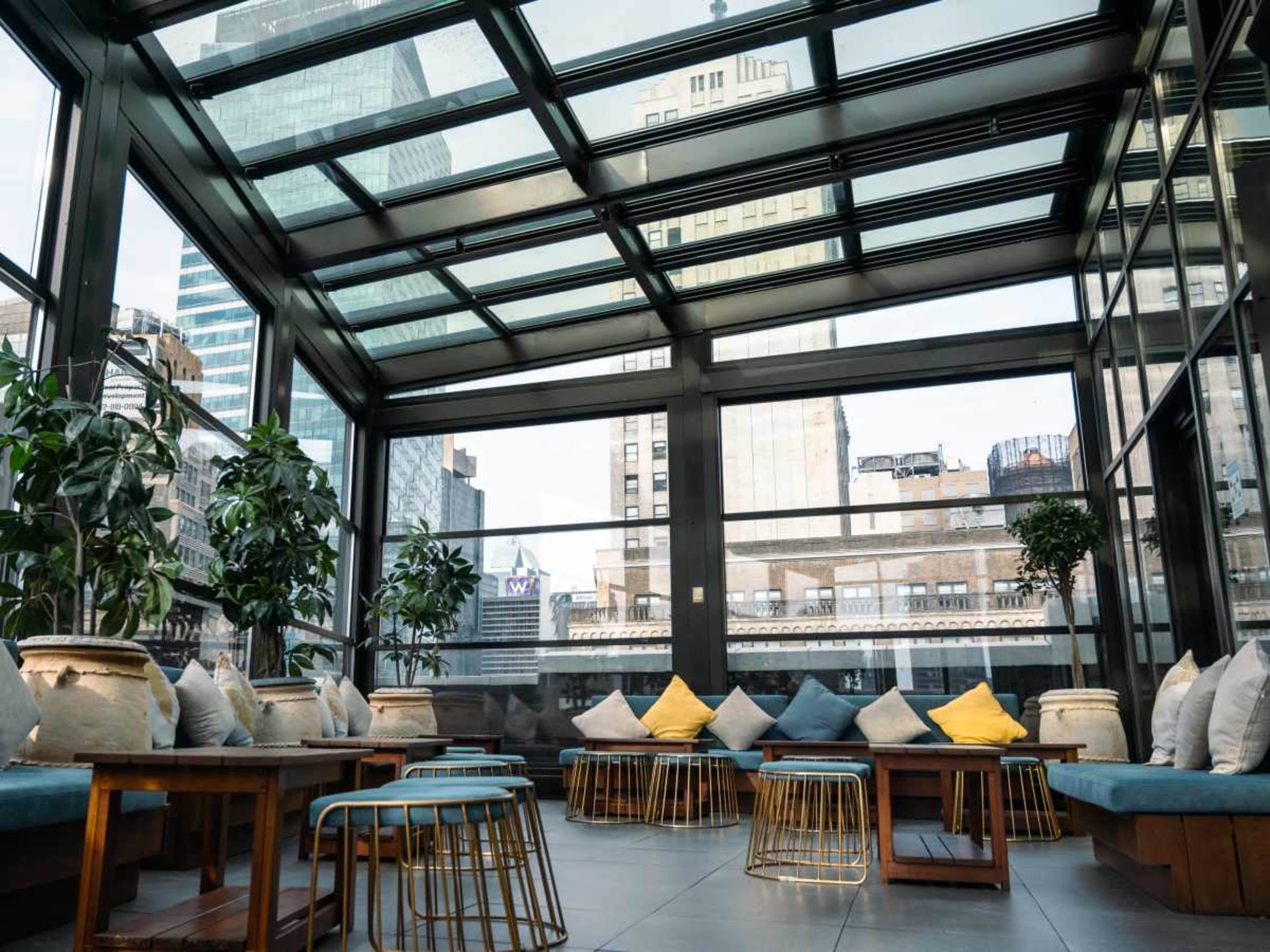 The image shows a modern indoor lounge area with glass walls, featuring wooden seating and potted plants, overlooking a city skyline.