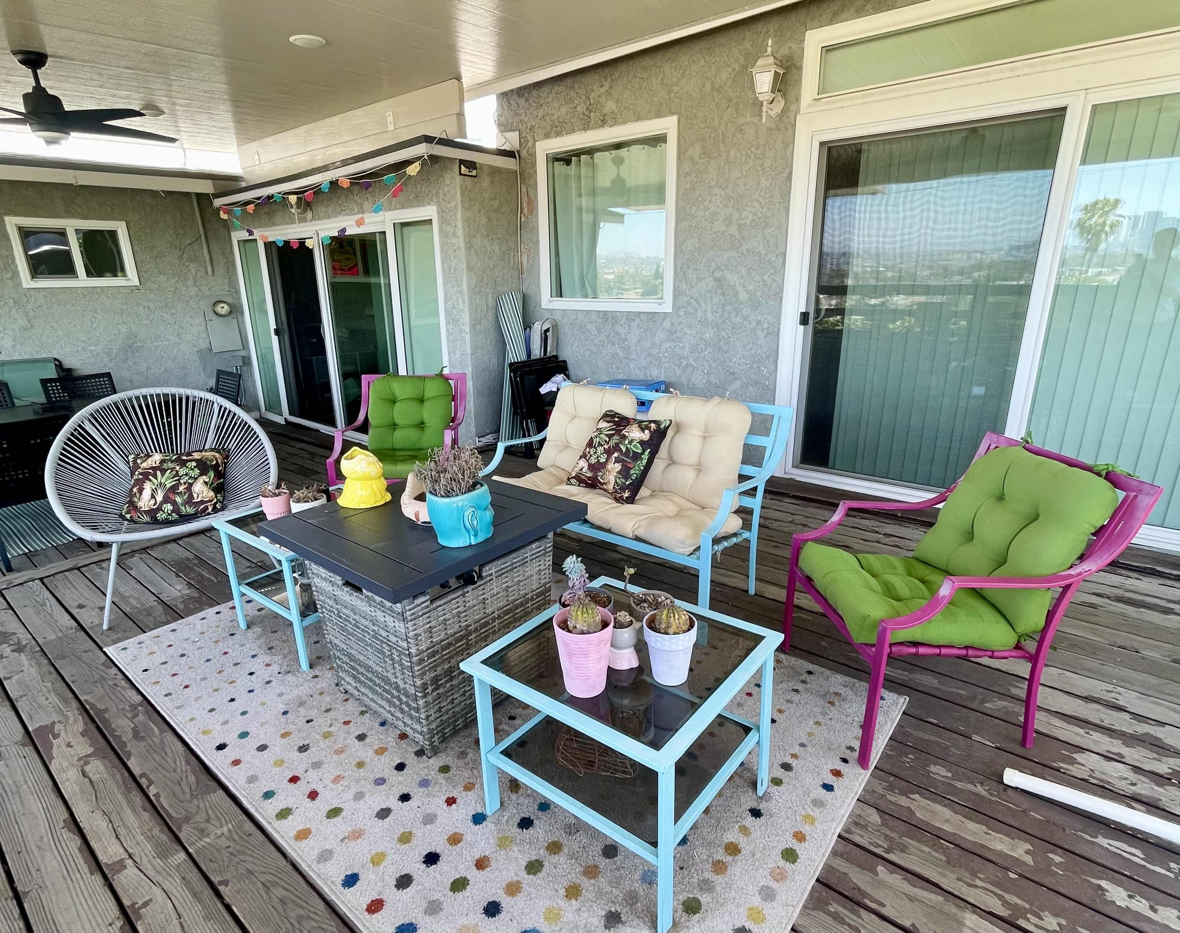 The image shows a patio furnished with colorful chairs, a coffee table, and potted plants on a wooden deck under a shaded roof.