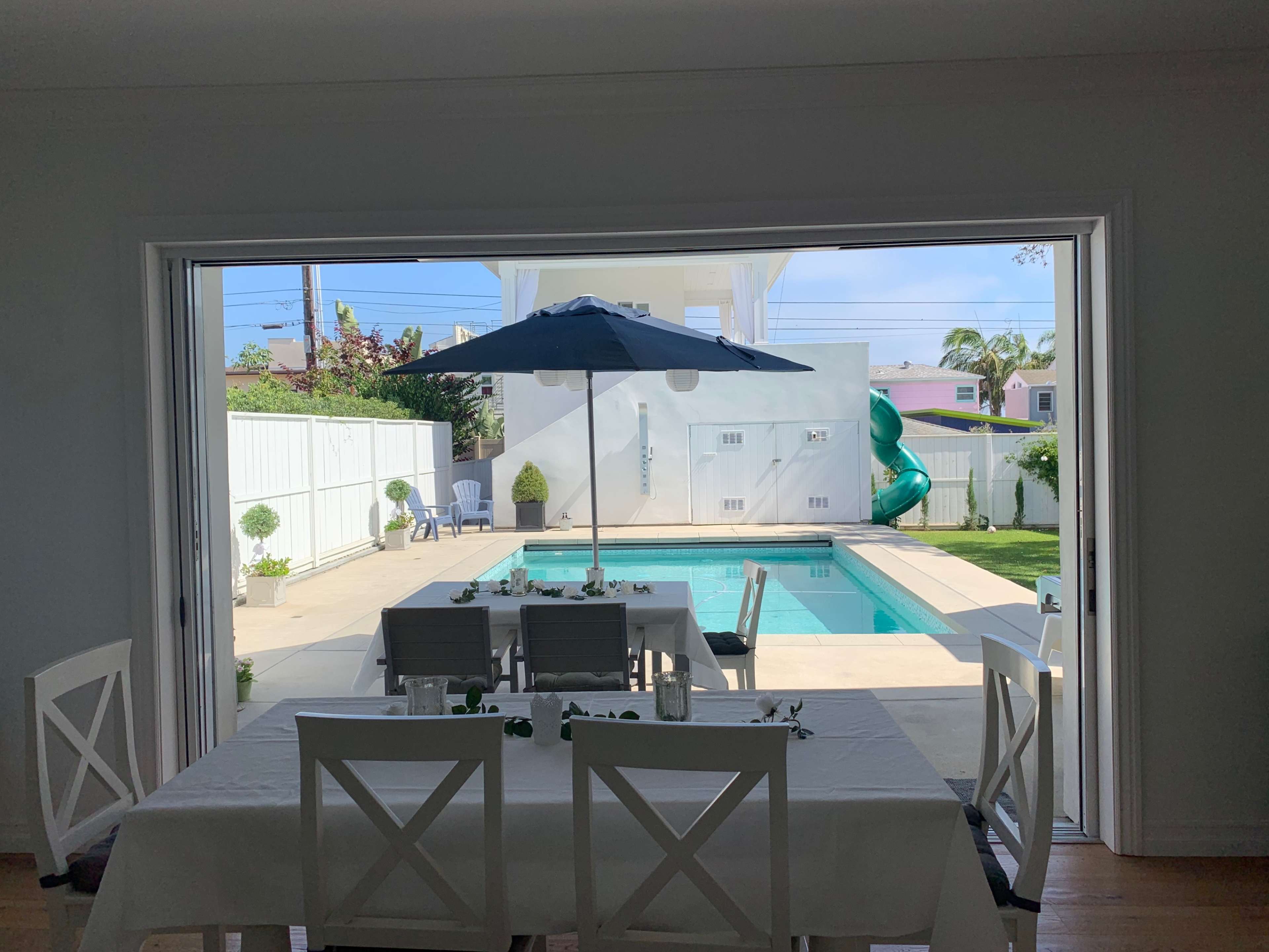 A dining area with a table under an umbrella overlooks a swimming pool and a backyard.