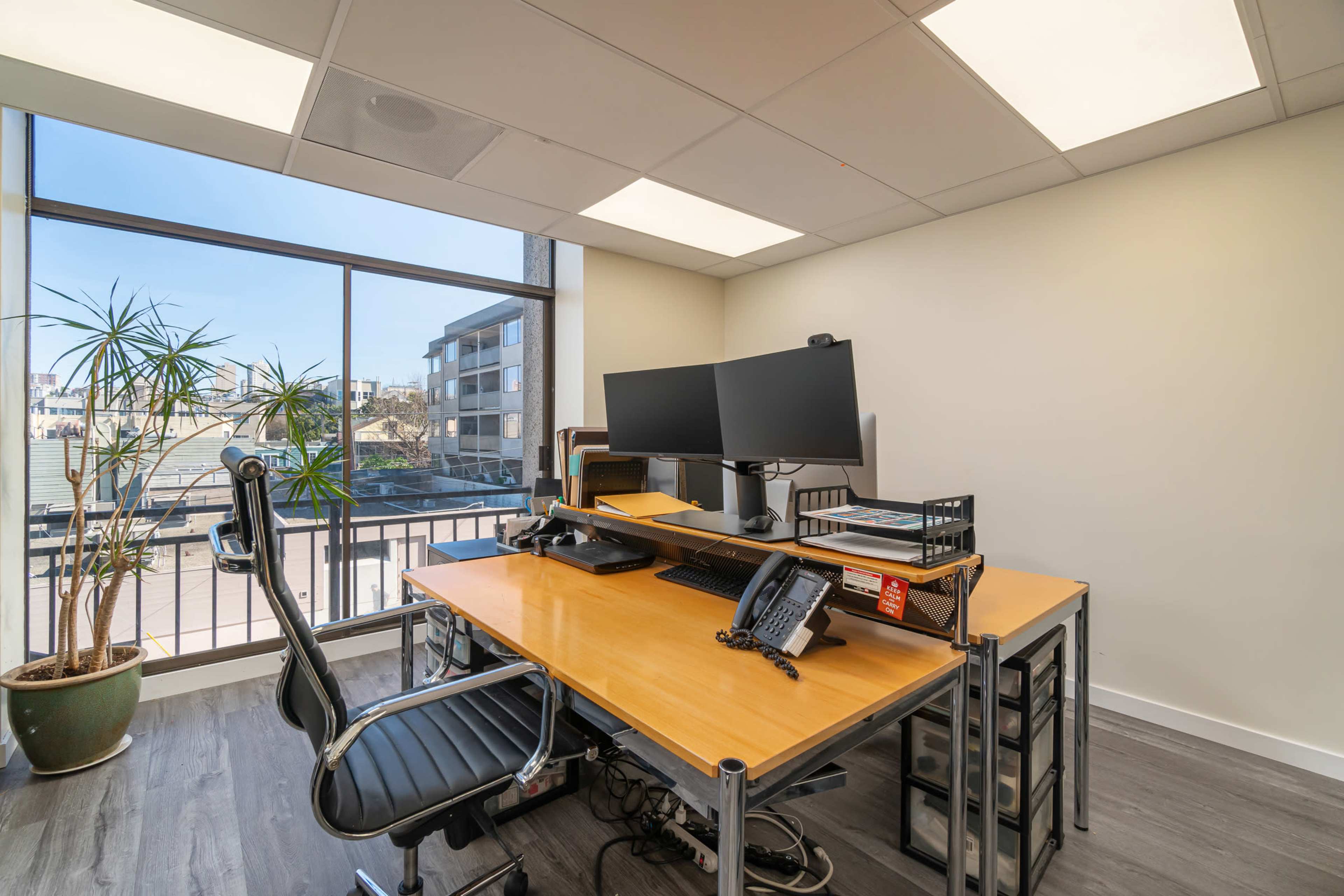 A spacious office features a wooden desk with dual monitors, an office chair, and a large window overlooking a cityscape.