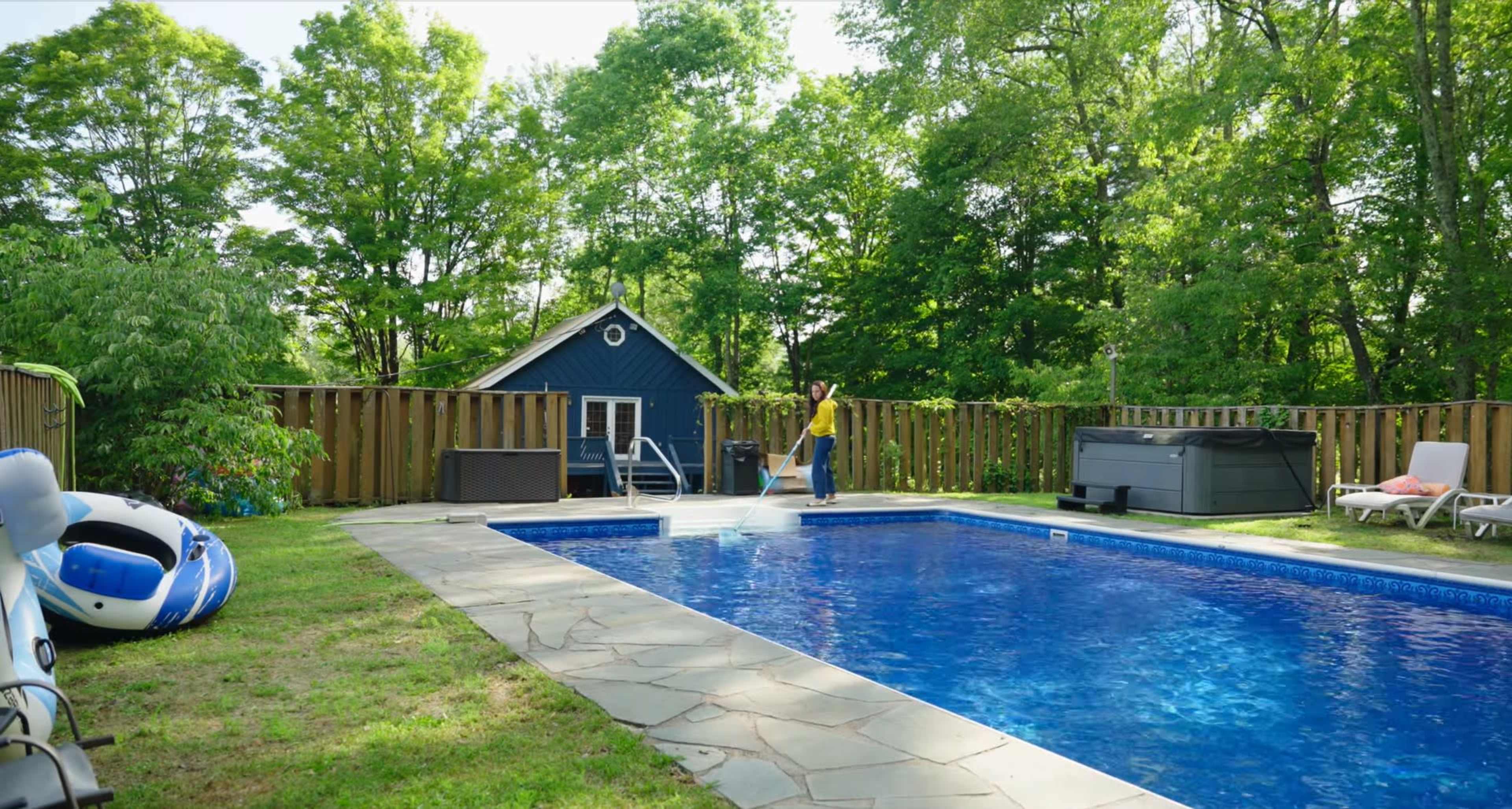 A person stands near a blue pool surrounded by green trees and a wooden fence, with a small blue building in the background.