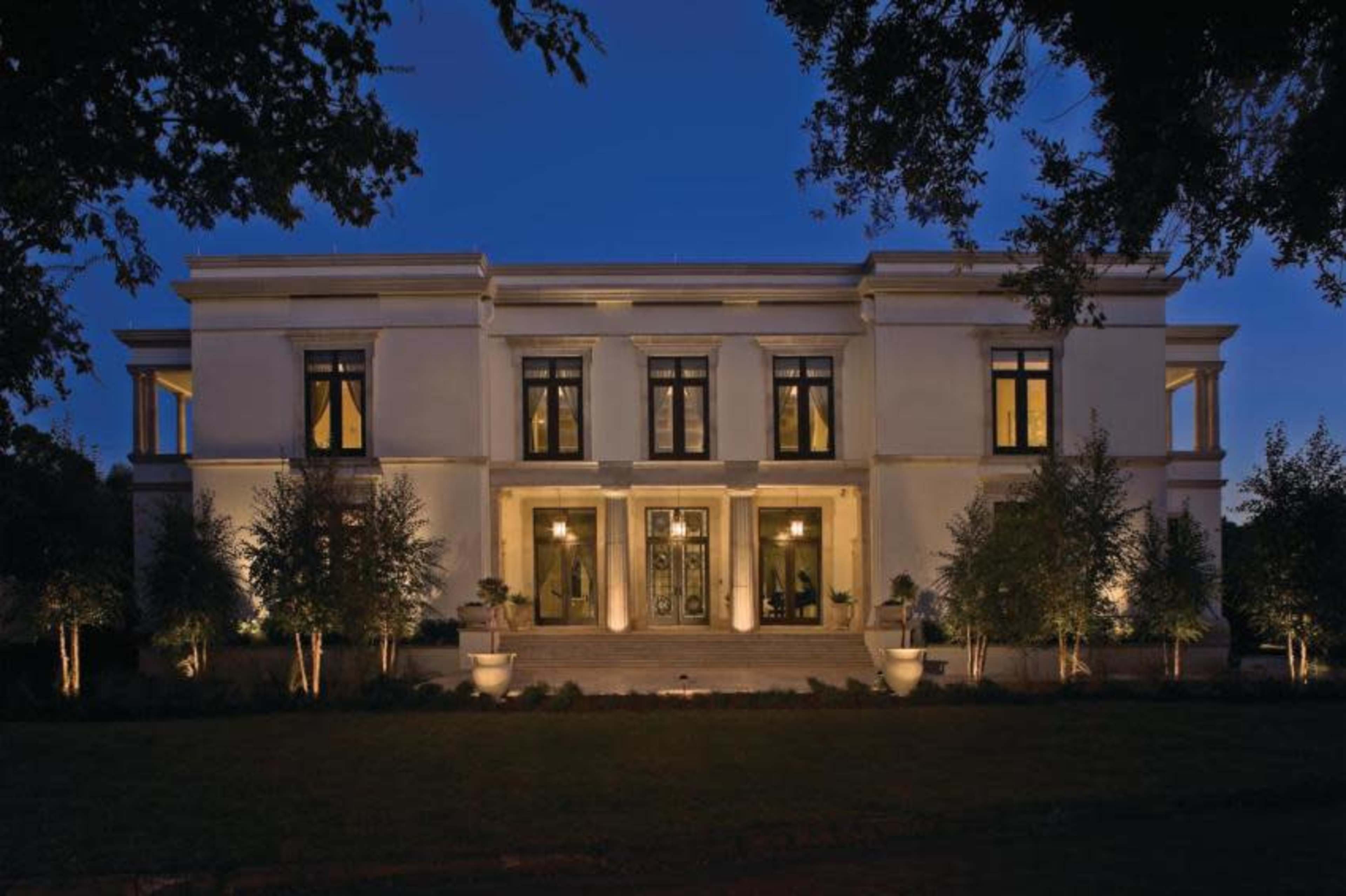 A modern, two-story white house with large windows and symmetrical features is illuminated by outdoor lighting at twilight.