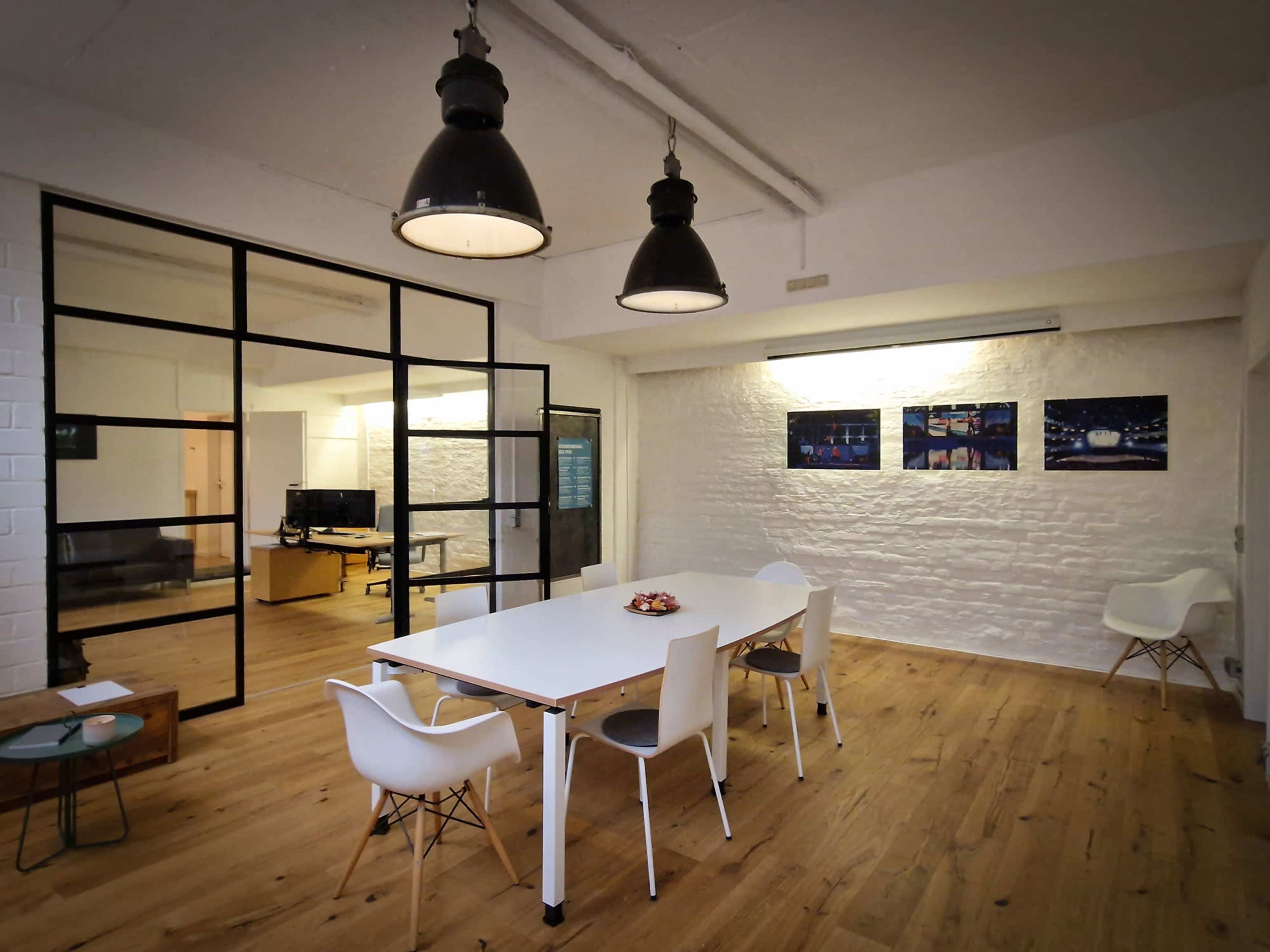 The image shows a modern office space featuring a white table surrounded by chairs, with a glass partition leading to another room and artwork displayed on the walls.