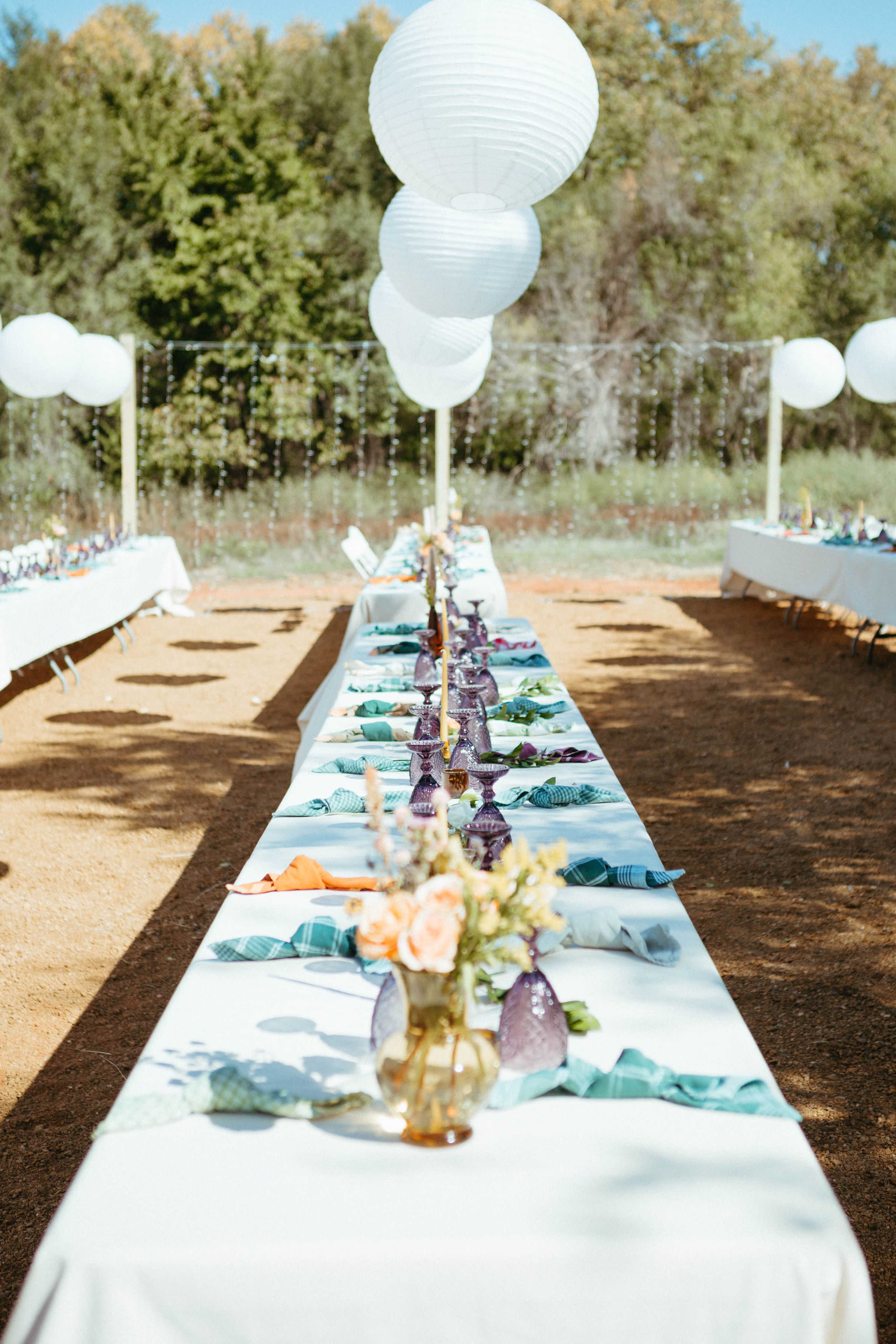 A long dining table is set outdoors, adorned with white tablecloths, colorful napkins, and vases of flowers, while oversized white lanterns hang above.