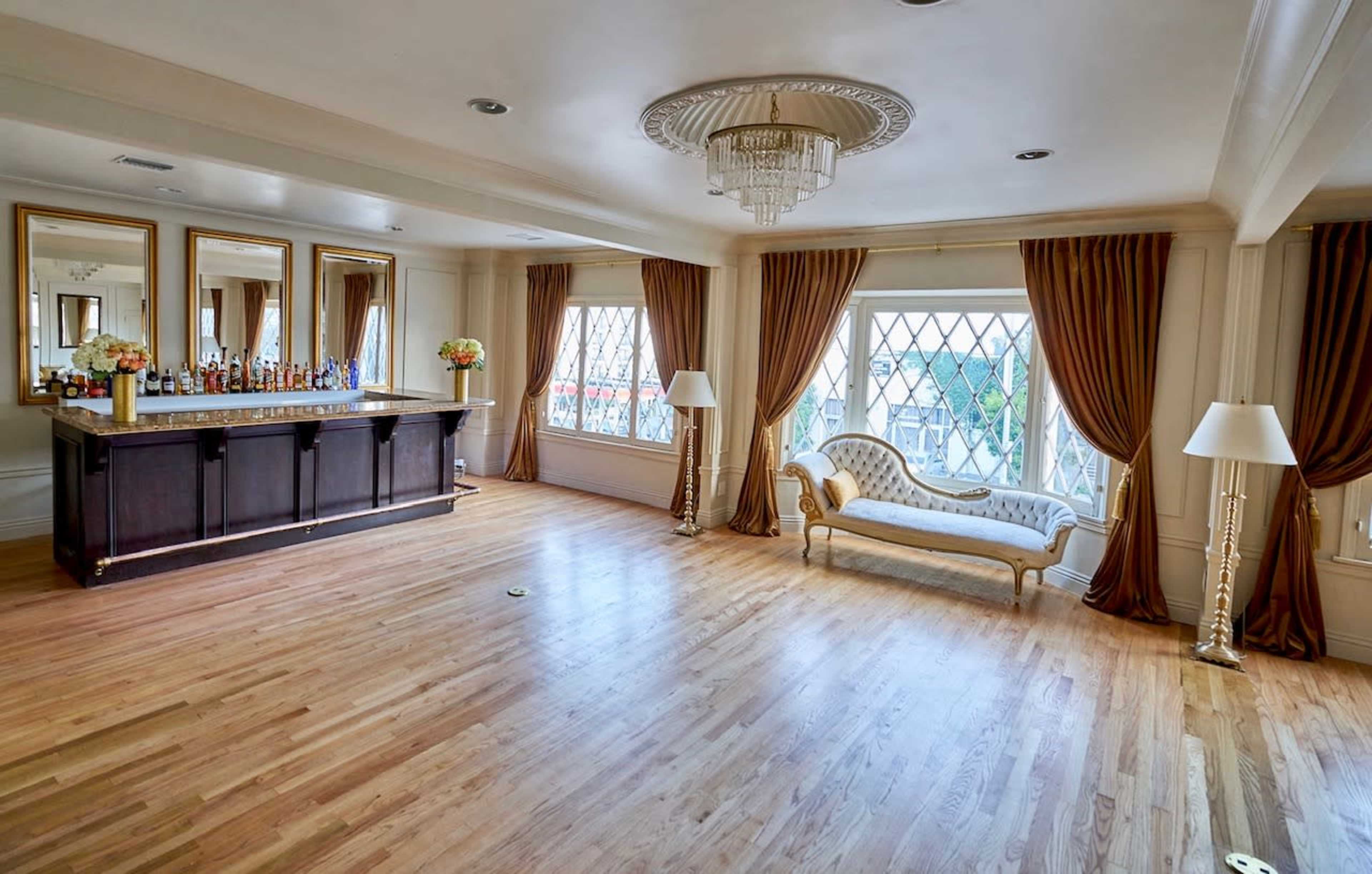 The image shows a spacious, elegantly designed room with a bar area, a sofa, and large windows framed by curtains.