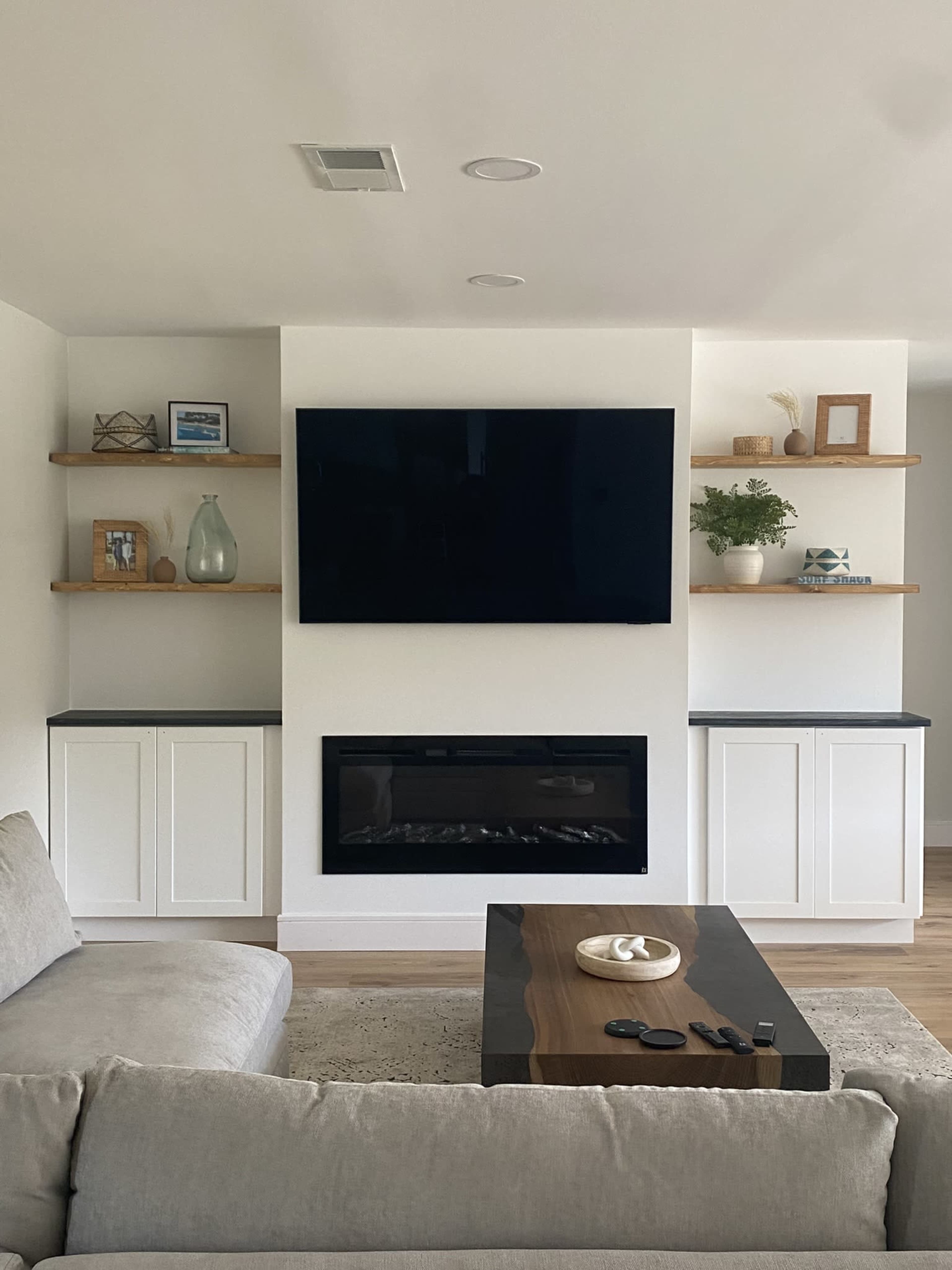 The image showcases a modern living room with a wall-mounted television, floating wooden shelves, a fireplace, and a light-colored sofa in the foreground.