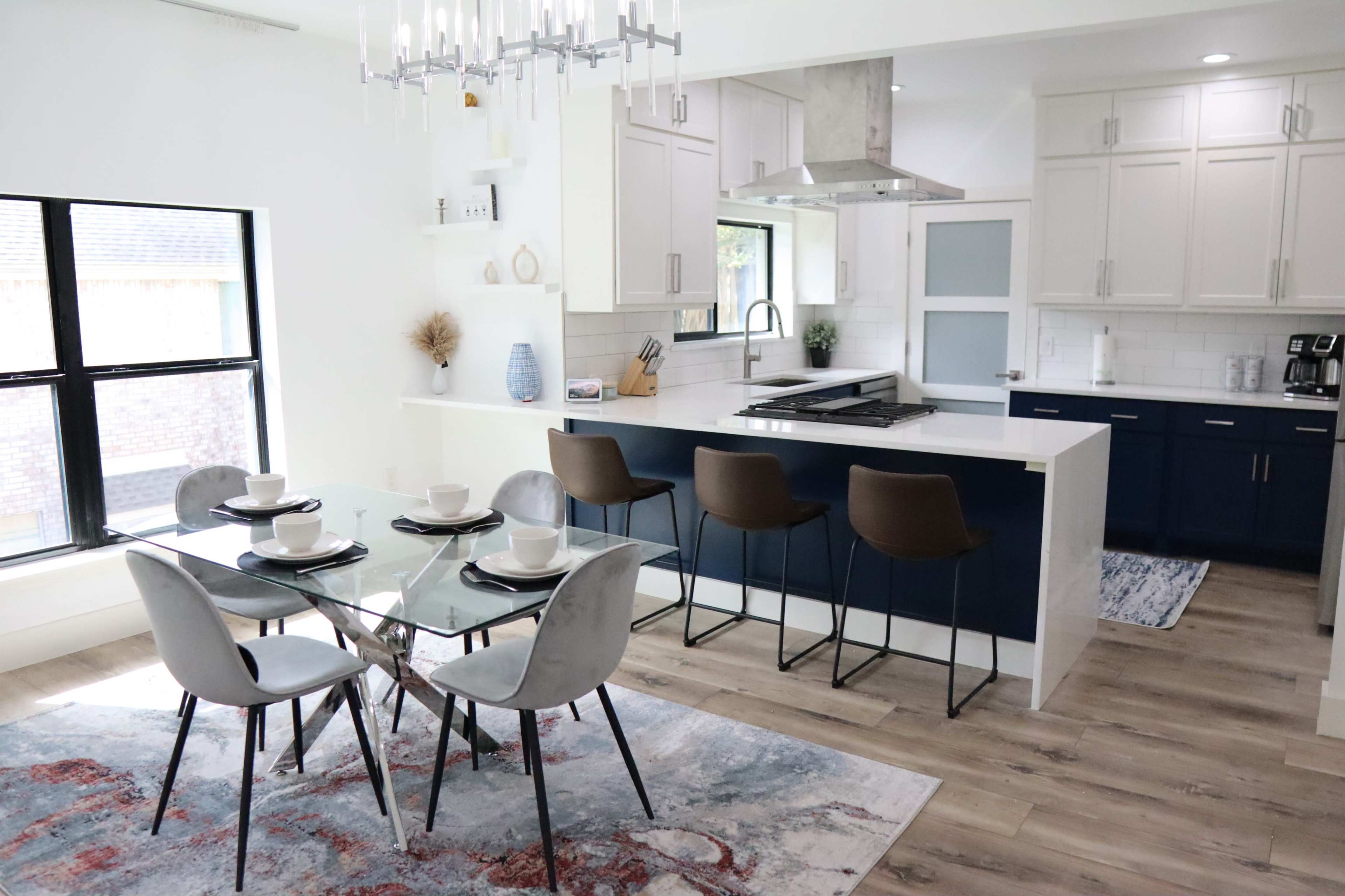 A modern kitchen and dining area feature a glass dining table with four chairs, a kitchen island with bar stools, and white cabinetry with navy blue accents.